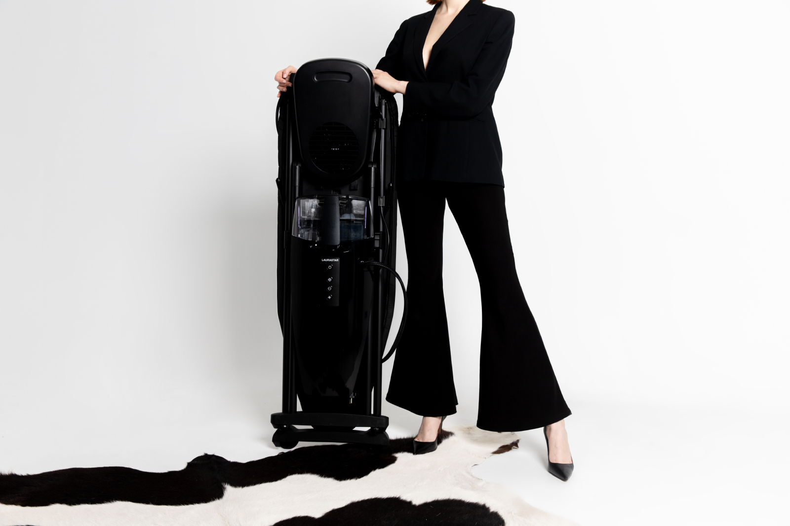 Kobieta w czarnym garniturze trzyma czarny parownik do odzieży, stojąc na czarno-białym dywanie ze skóry krowy.