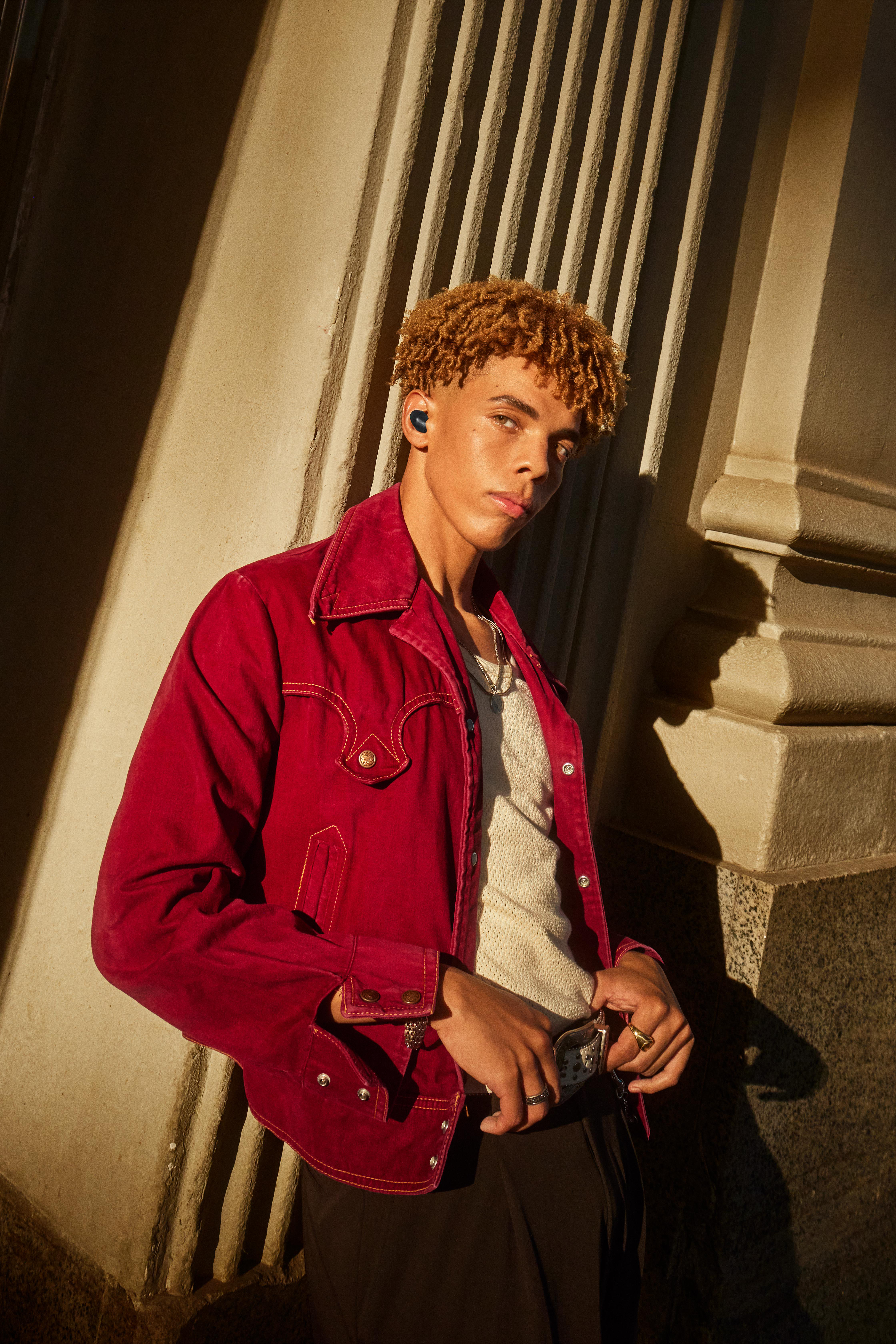 Junger Mann in roter Jacke, lehnt an Gebäude, trägt Kopfhörer. Sonniger Beleuchtung.