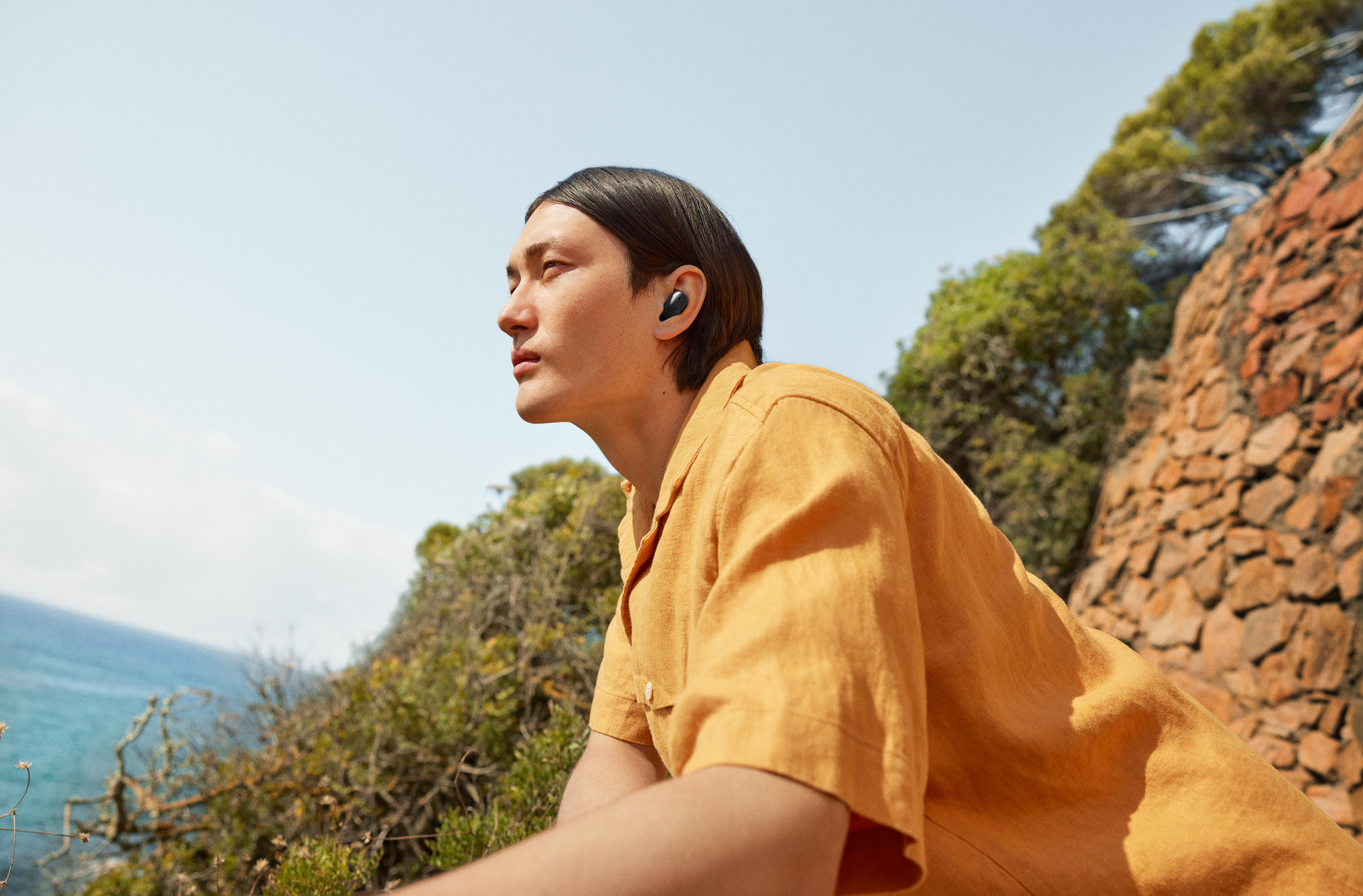 Ein Mann schaut nach oben, trägt ein orangefarbenes Hemd, Ohrhörer, nahe grünen Büschen und dem Meer.