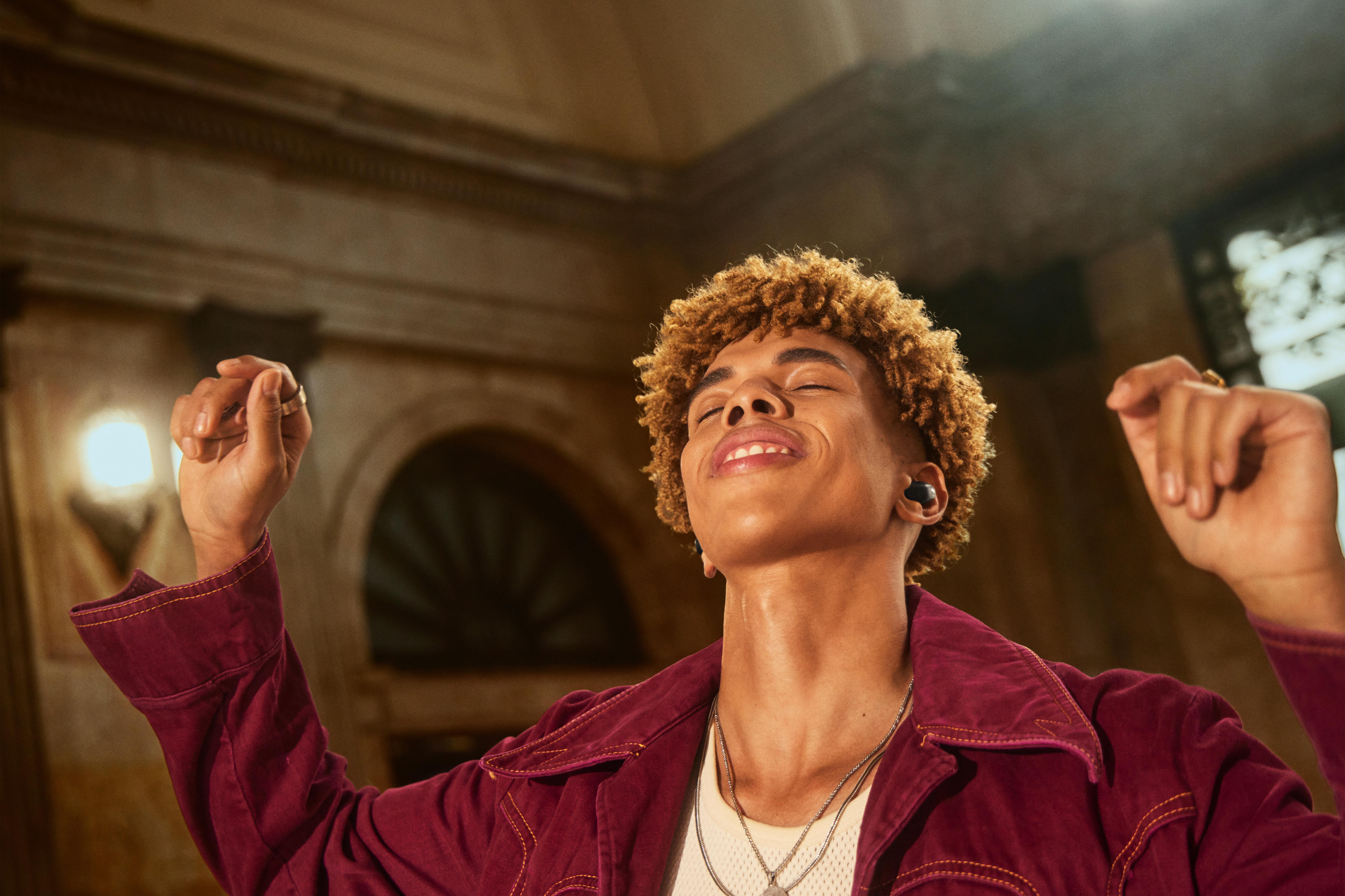 Ein junger Mann mit lockigem Haar, der eine rote Jacke trägt und mit Ohrhörern Musik hört.