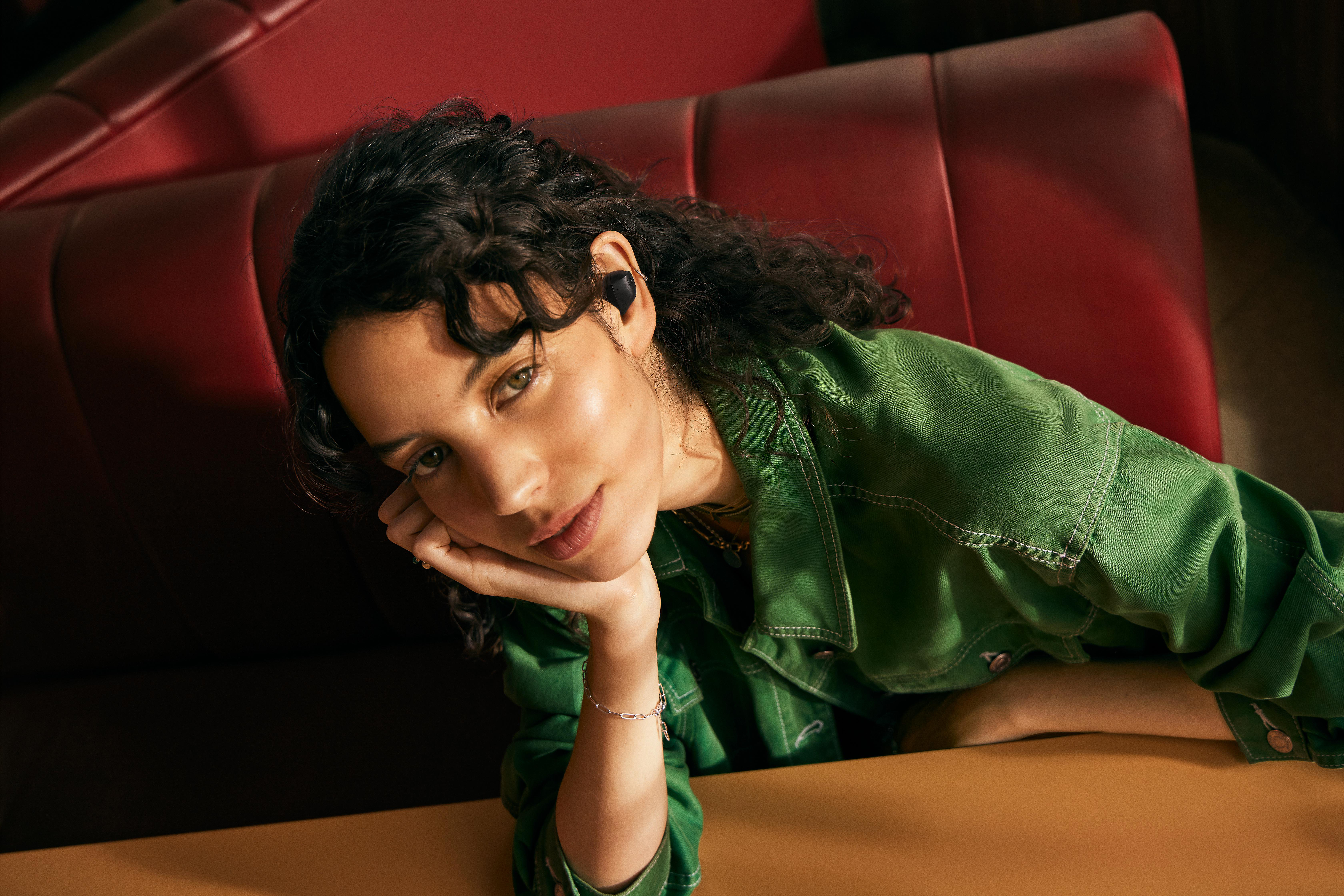 Frau mit dunklen Haaren, grüne Jacke, Ohrhörer. Lehnt an einem Tisch.