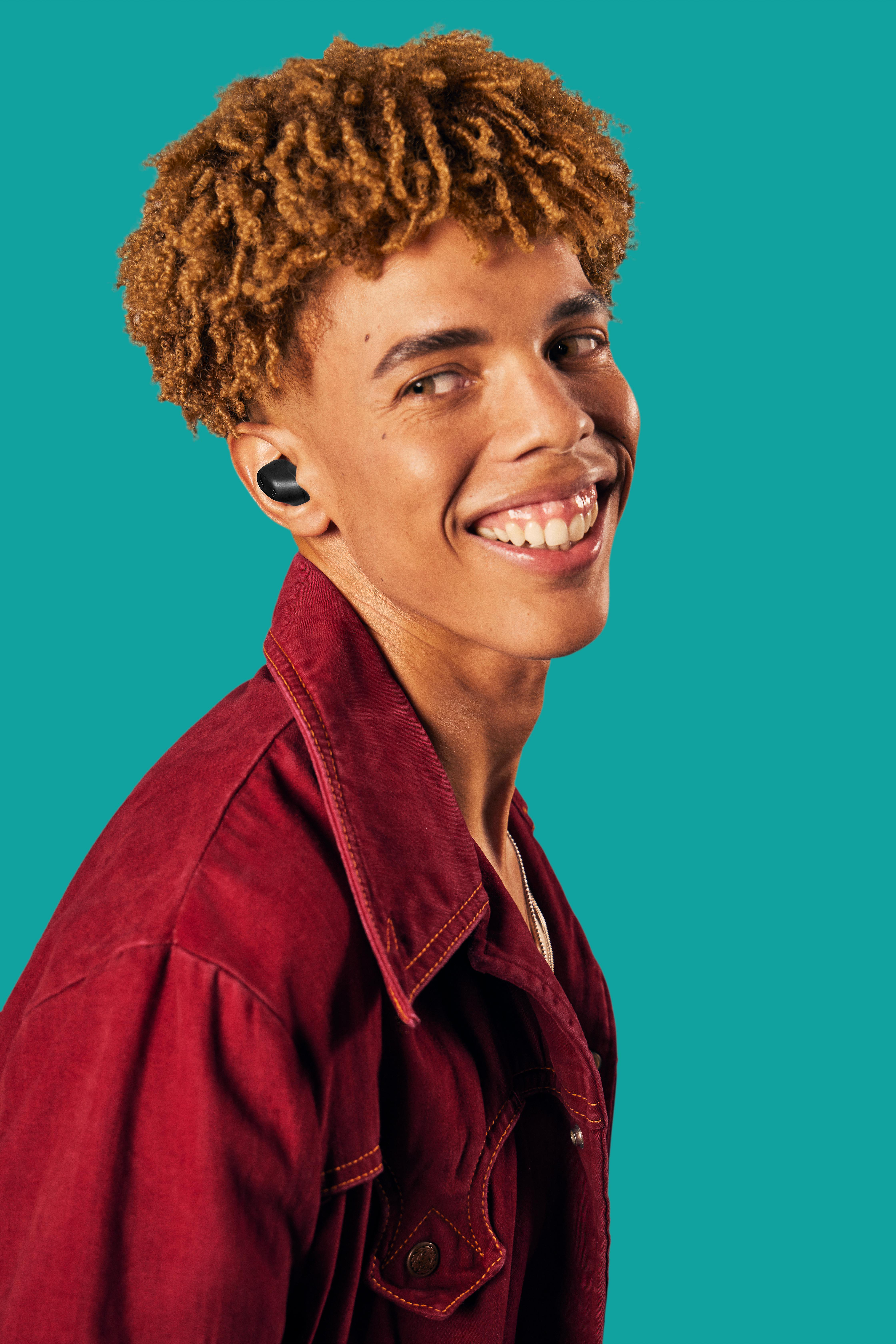 Lächelnder junger Mann mit lockigem Haar, der Ohrhörer und eine rote Jacke vor teal Hintergrund trägt.