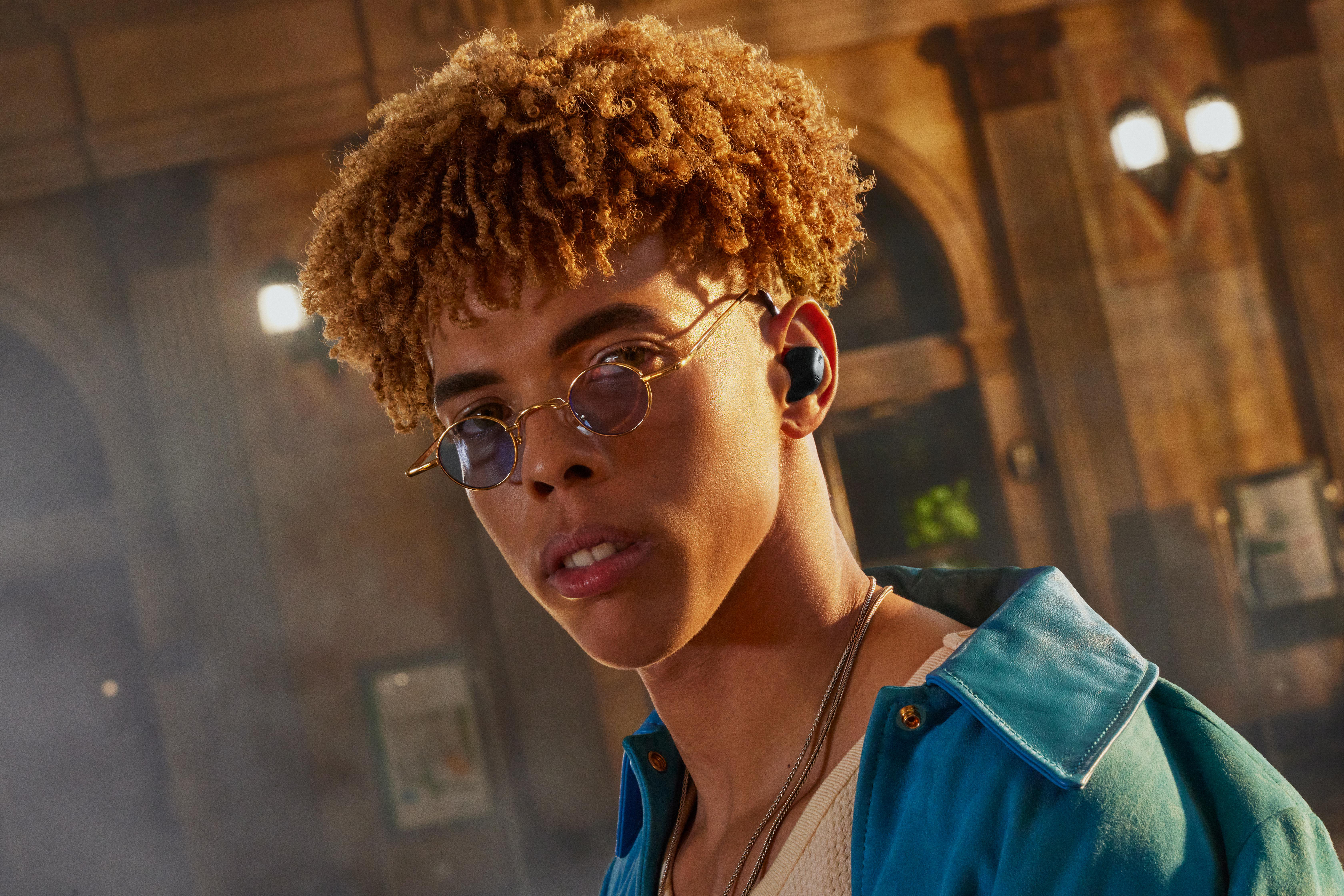 Ein junger Mann mit lockigem Haar, Brille und Ohrhörern.