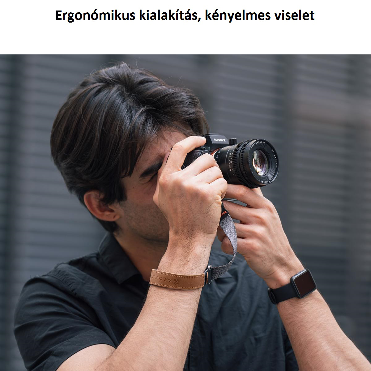 Egy férfi kamerát tart, fényképez. Fekete inget és órát visel. A kamerán lencse és szíj van.