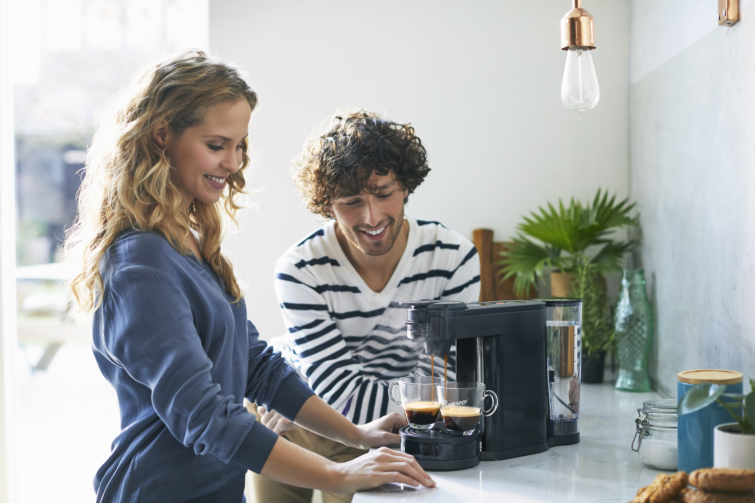 Paar lächelt in einer Küche und bereitet Kaffee mit einer schwarzen Kaffeemaschine zu.