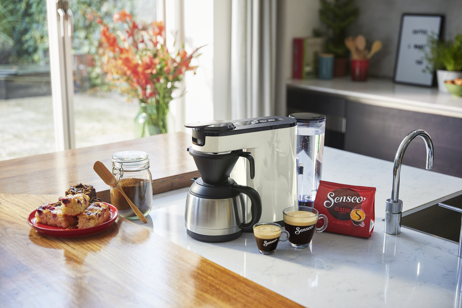 Koffiezetapparaat met koffiekopjes, bonen en een bord gebak op een aanrecht.