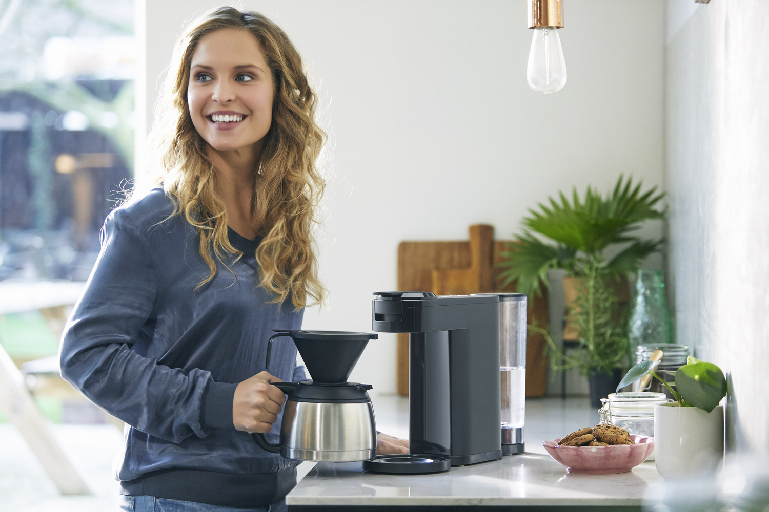 Een vrouw in een blauwe jas glimlacht, met een koffiepot in haar hand. Een zwart koffiezetapparaat en planten zijn zichtbaar.