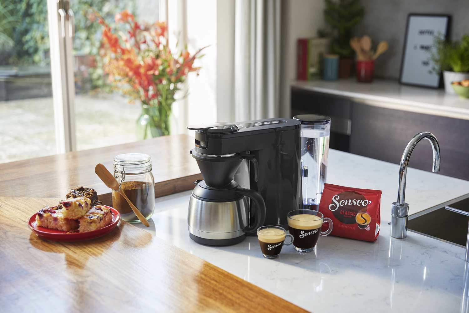 Koffiezetapparaat met koffie, gebak en ingrediënten op een aanrecht in de keuken, bij een raam.