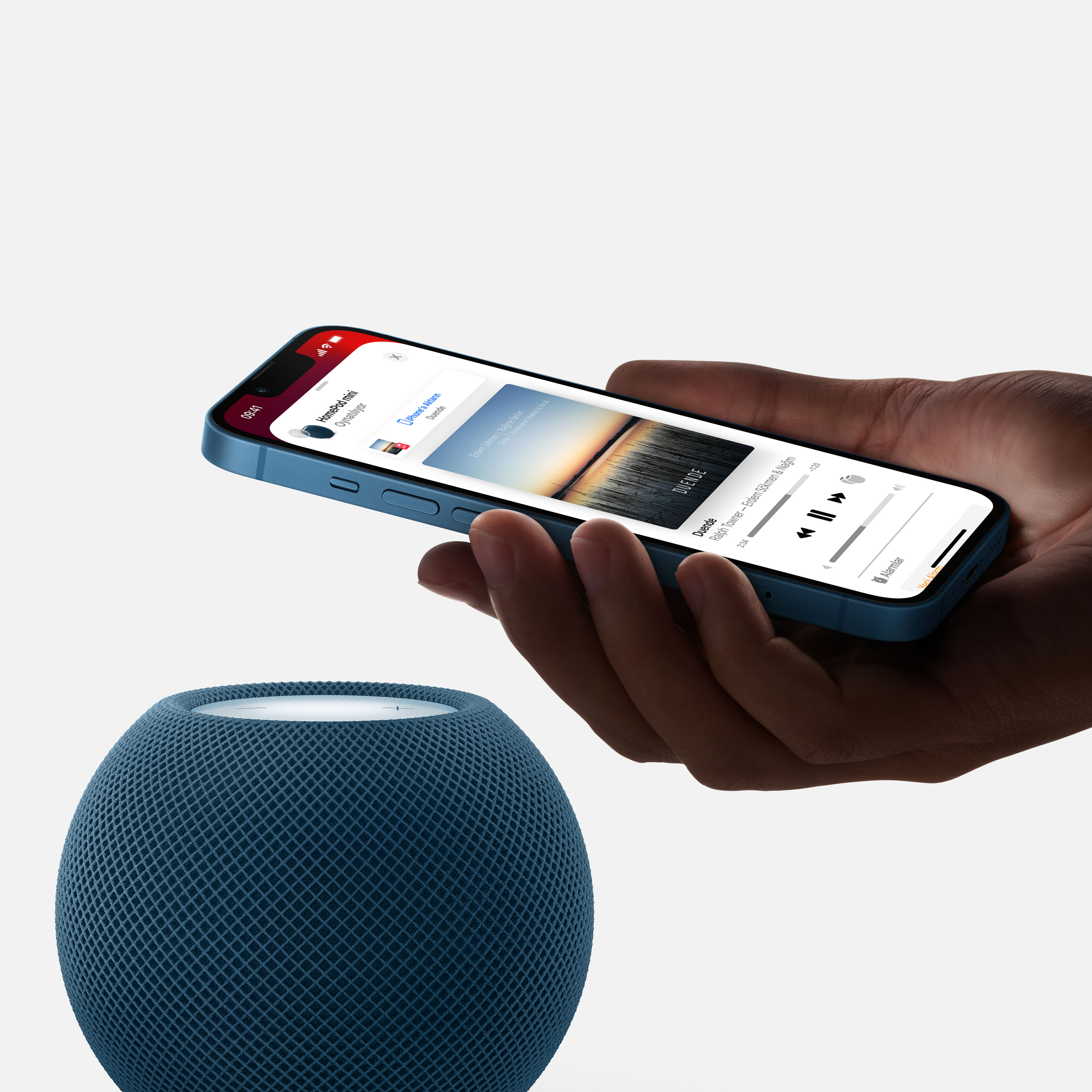 Müzik uygulaması arayüzünü gösteren mavi bir iPhone tutan bir kişi. Masada mavi bir HomePod Mini var.