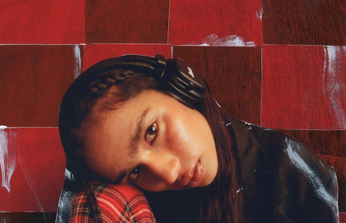 Osoba przed czerwoną i brązową ścianą ma na sobie słuchawki i patrzy w kierunku kamery.