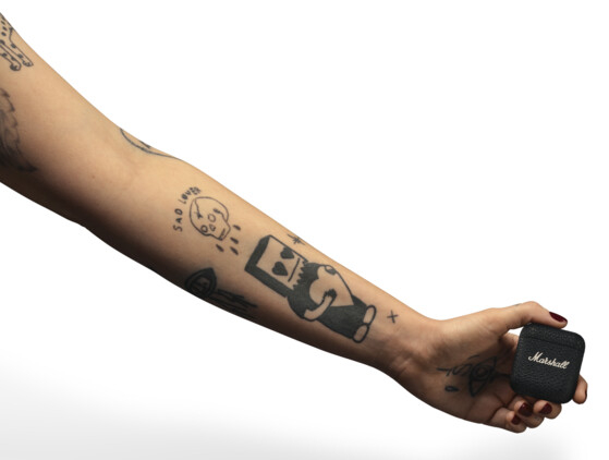 Osoba z tatuażami trzyma małą, czarną obudowę głośnika Marshall. Obudowa ma biały tekst.