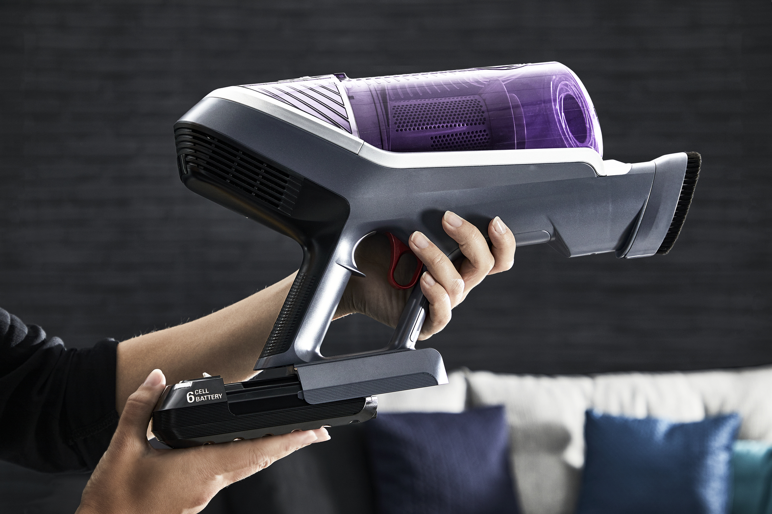 A hand holding a black and gray cordless vacuum cleaner. The dustbin is purple.