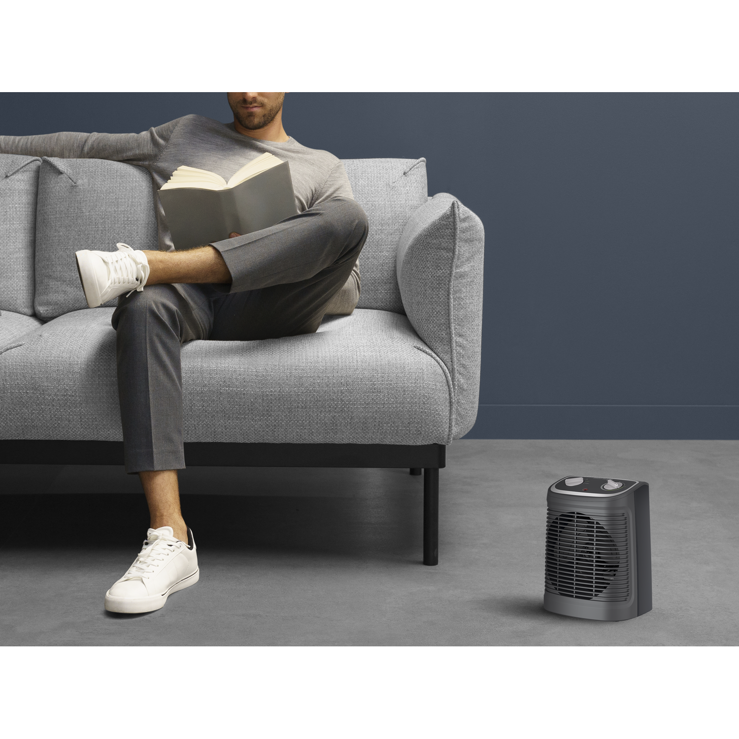 A man sitting on a gray couch with a book, a black heater nearby.