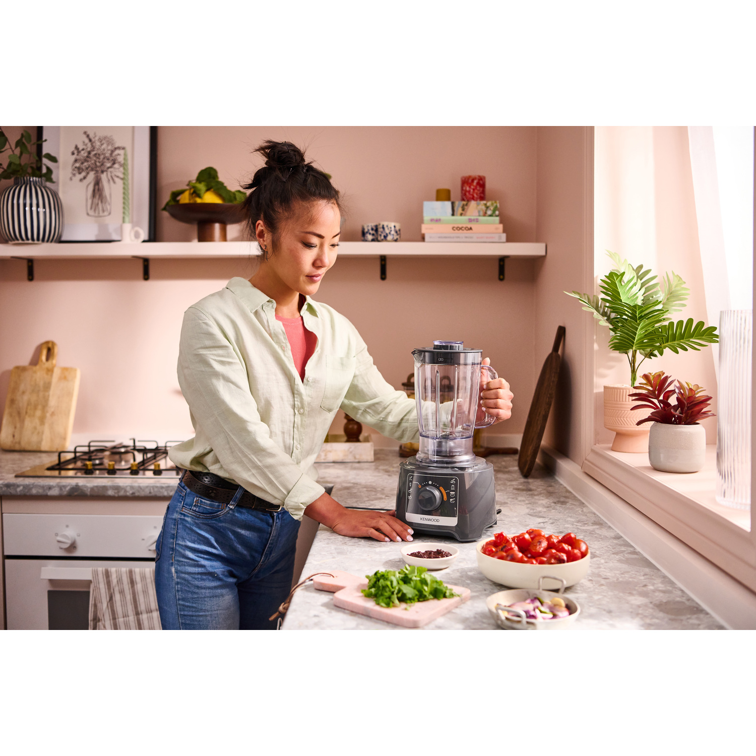 Une femme prépare de la nourriture avec un mixeur sur un comptoir de cuisine. Des ingrédients comme des tomates sont visibles.