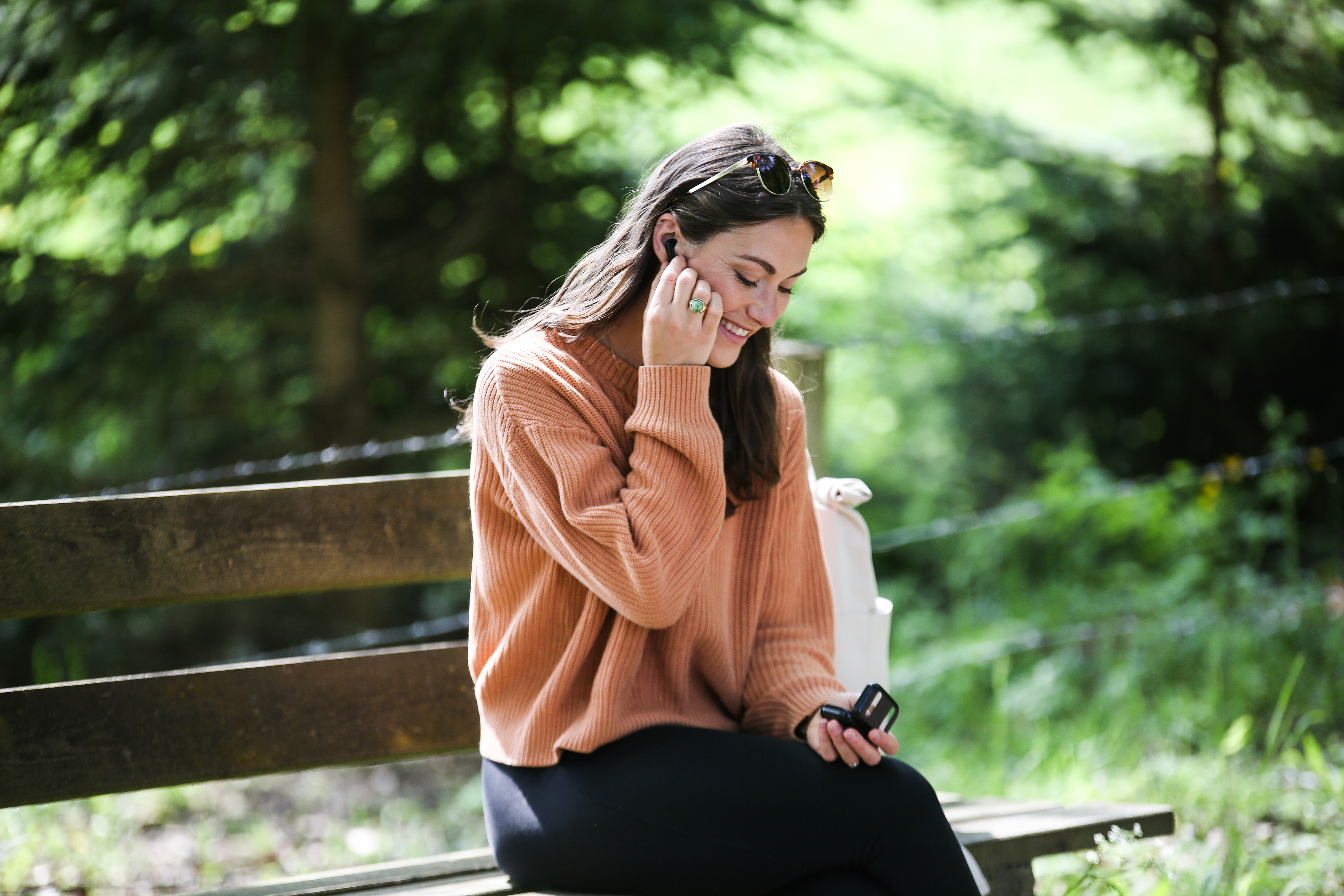 Kobieta w pomarańczowym swetrze i okularach na drewnianej ławce, używa słuchawek. Tło to zielone drzewa.