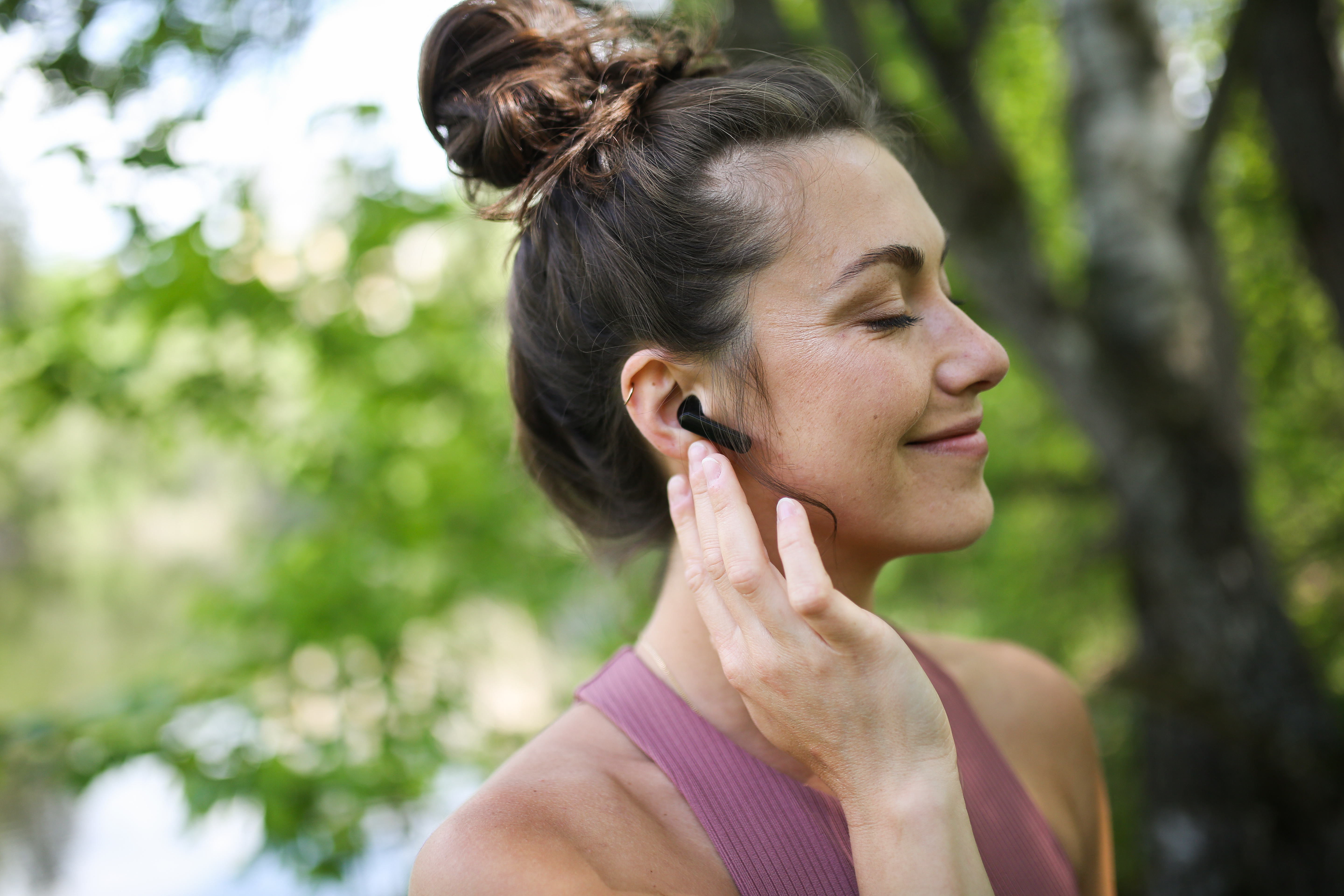 Kobieta w słuchawkach w plenerze, uśmiechnięta i dotykająca ucha.