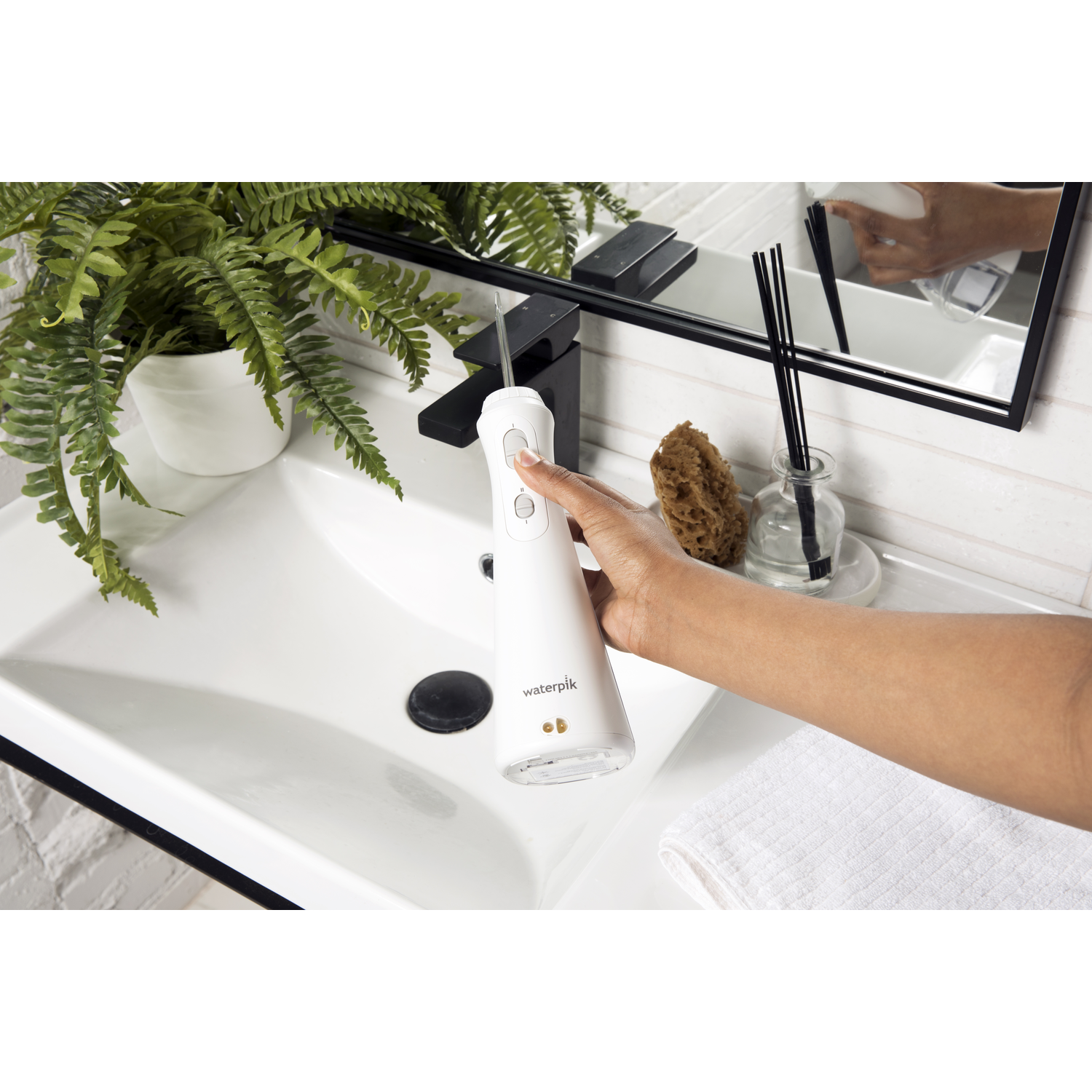 Een persoon die een Waterpik-monddouche gebruikt boven een witte wastafel met spiegel en accessoires.