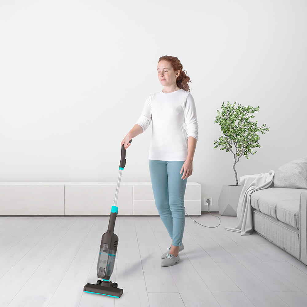 Kobieta odkurza pokój z białą ścianą, małym drzewkiem, sofą i białą szafką w tle.