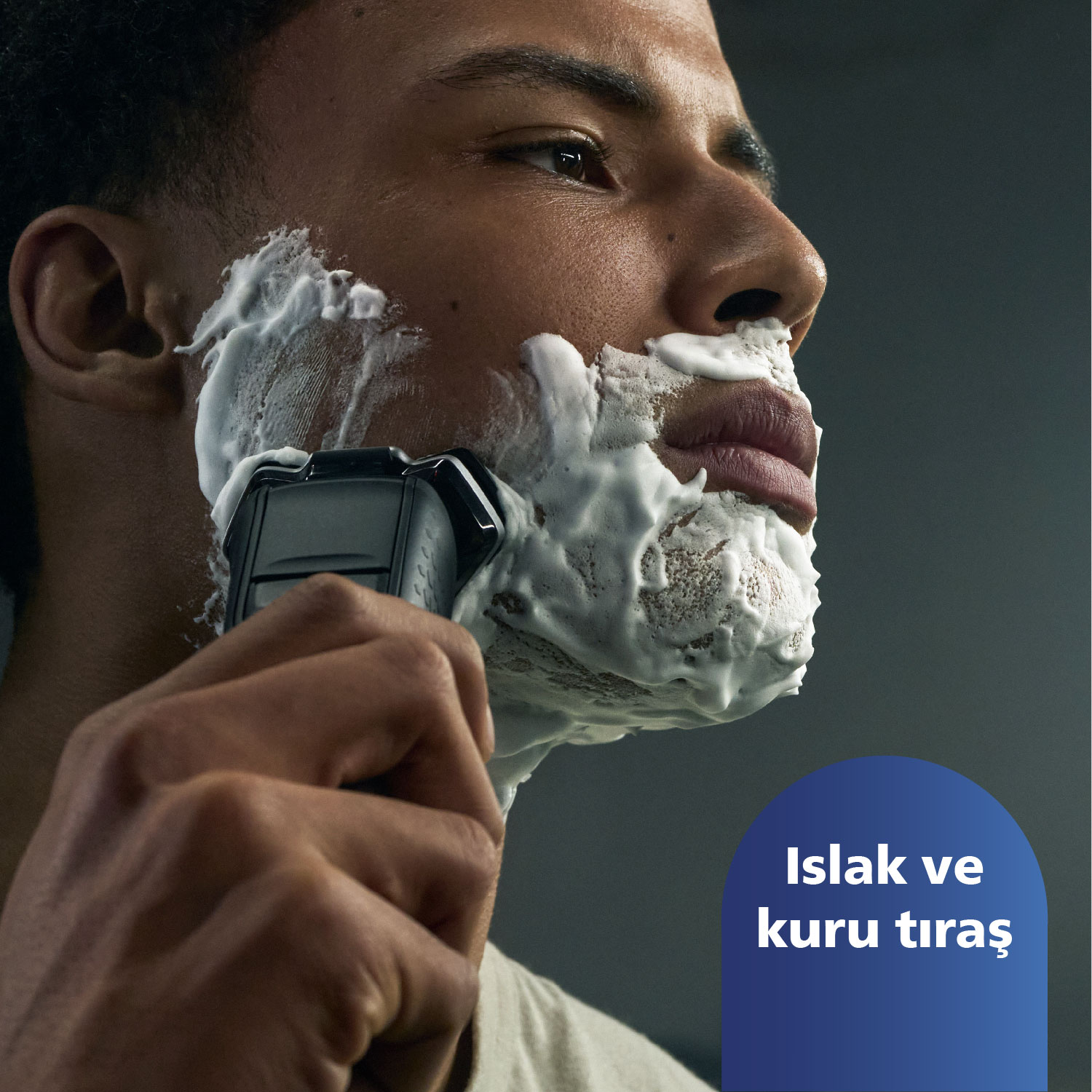 Erkek elektrikli tıraş makinesiyle tıraş oluyor. Yüzünde köpük. Karanlık arka plan.