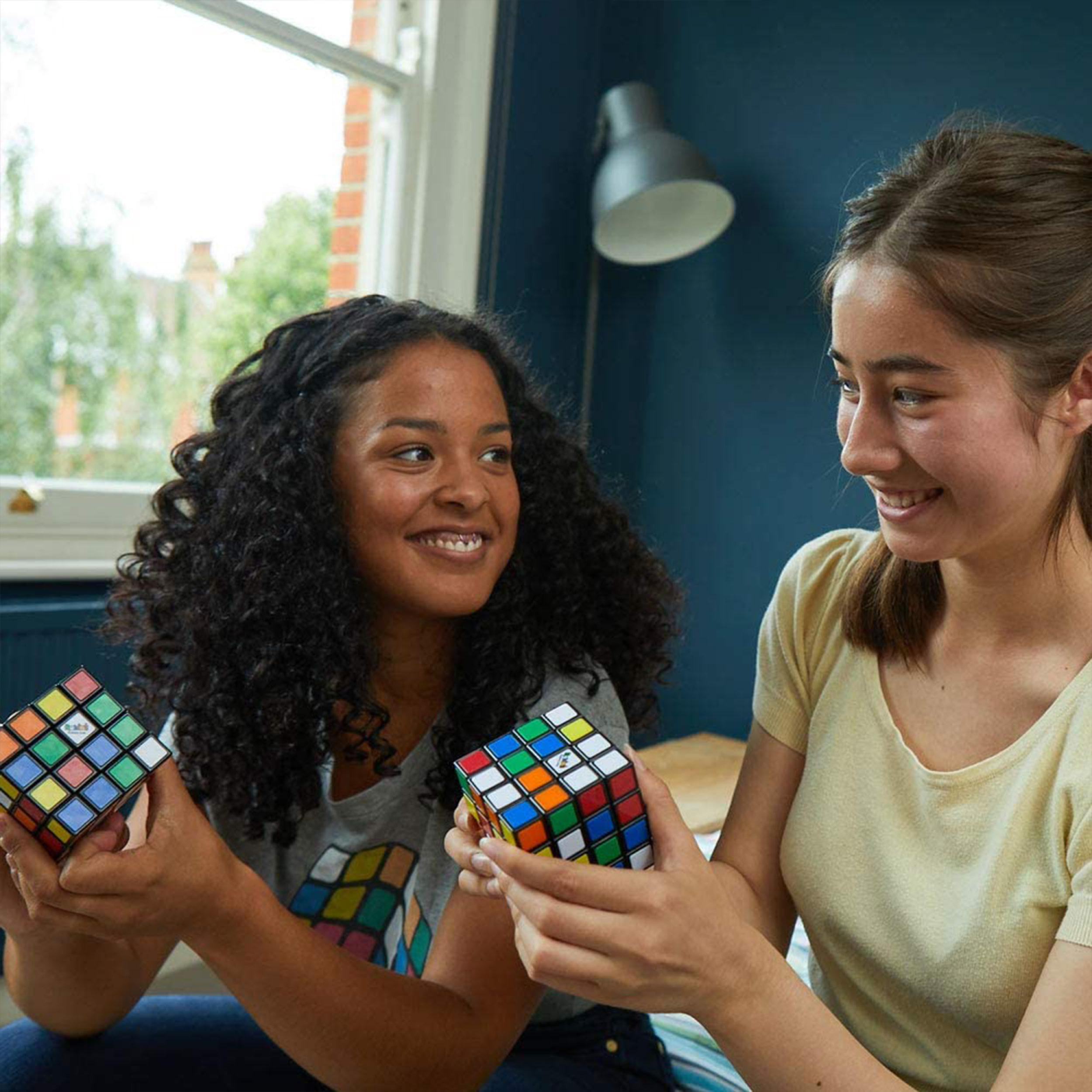 Zwei junge Frauen halten Rubik's Würfel, lächeln. Hintergrund ist ein Fenster und eine Wand.