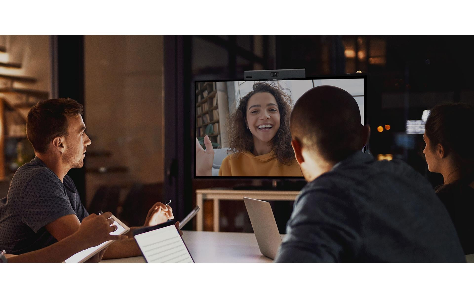 Drei Personen an einem Tisch betrachten einen Monitor mit einer Frau bei einem Videoanruf.