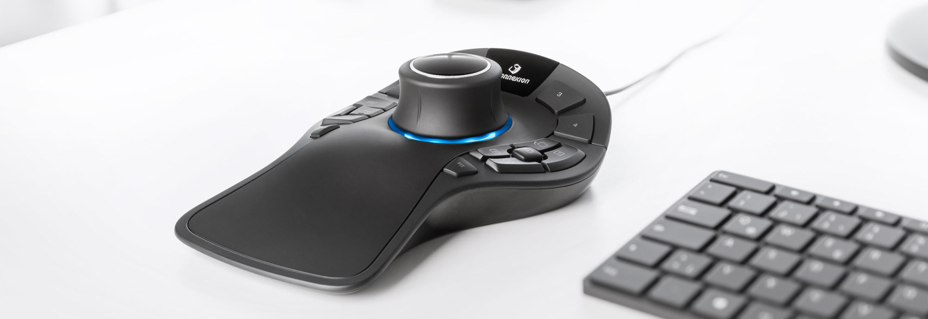 Un mouse 3Dconnexion nero e grigio è mostrato su una scrivania bianca, accanto a una tastiera. Il mouse ha dei pulsanti.