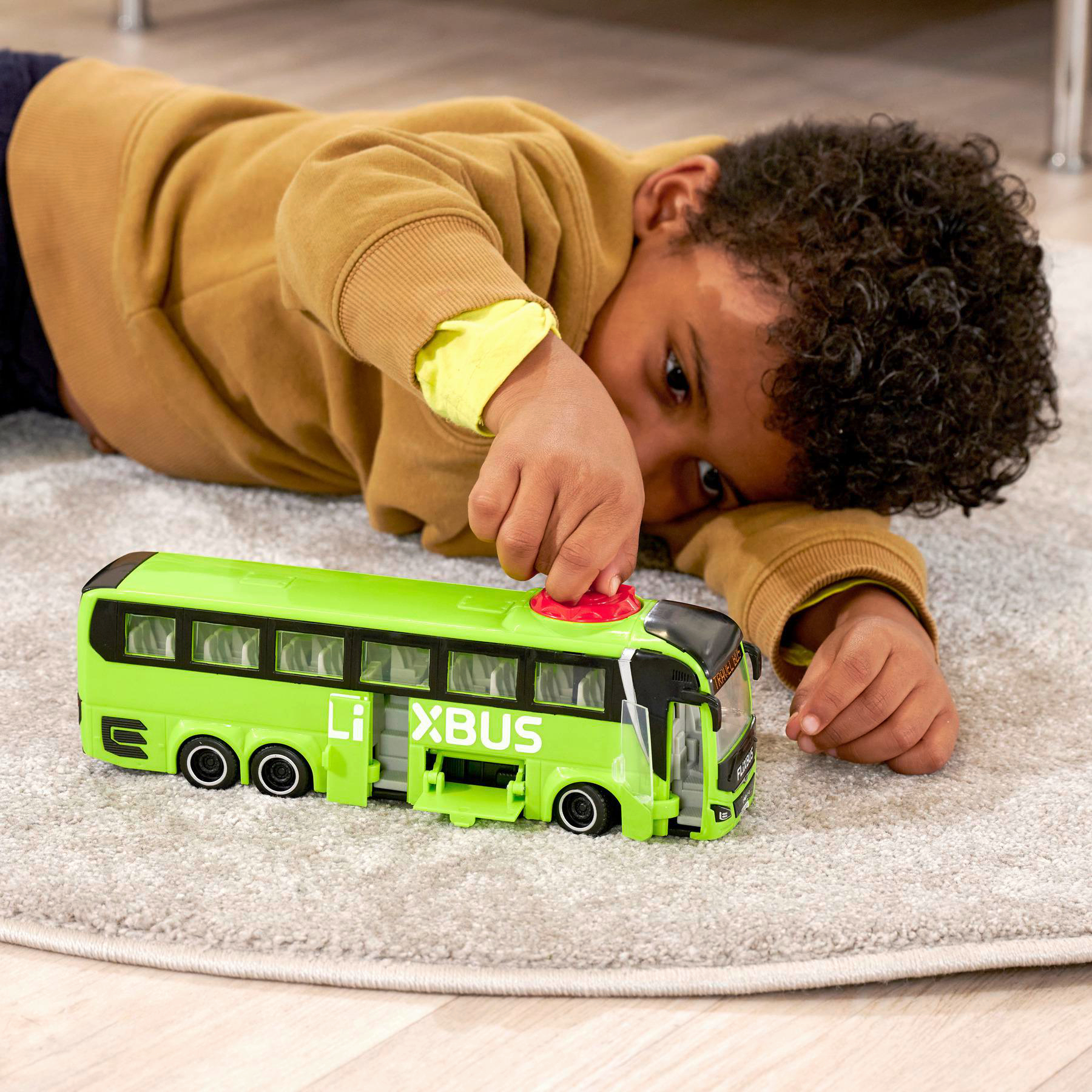 Ein kleiner Junge liegt auf dem Boden und spielt mit einem grünen Spielzeugbus. Der Bus hat schwarze Fenster und Räder.