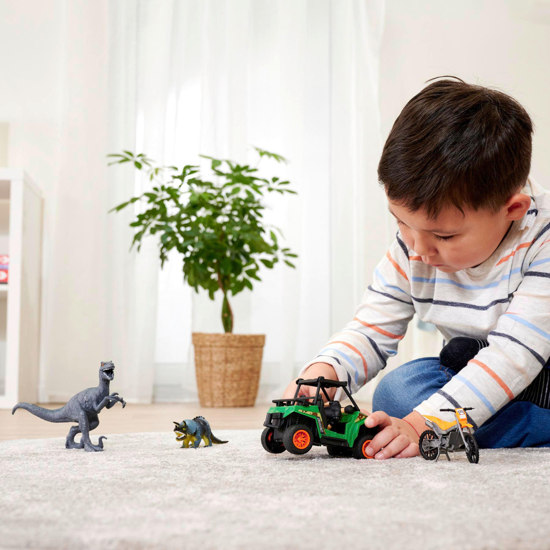 Junge spielt mit Spielzeugdinosauriern, grünem ATV und Motorrad auf einem Teppich.