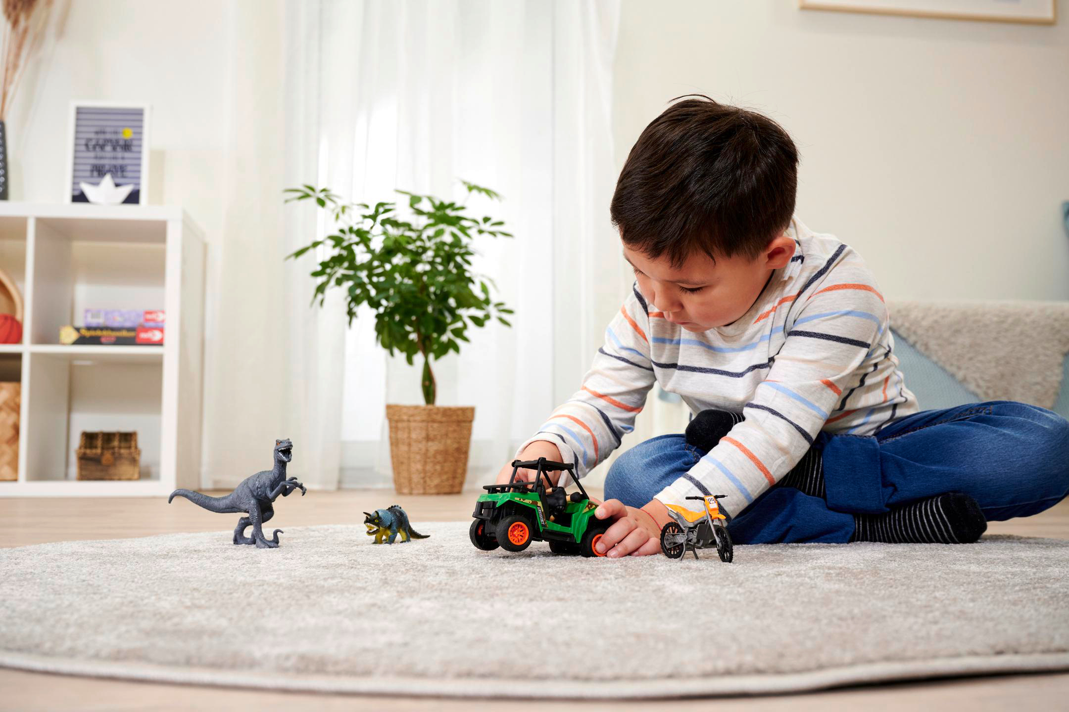Ein Kind spielt mit Spielzeugdinosauriern und einem grünen Jeep auf grauem Teppich.
