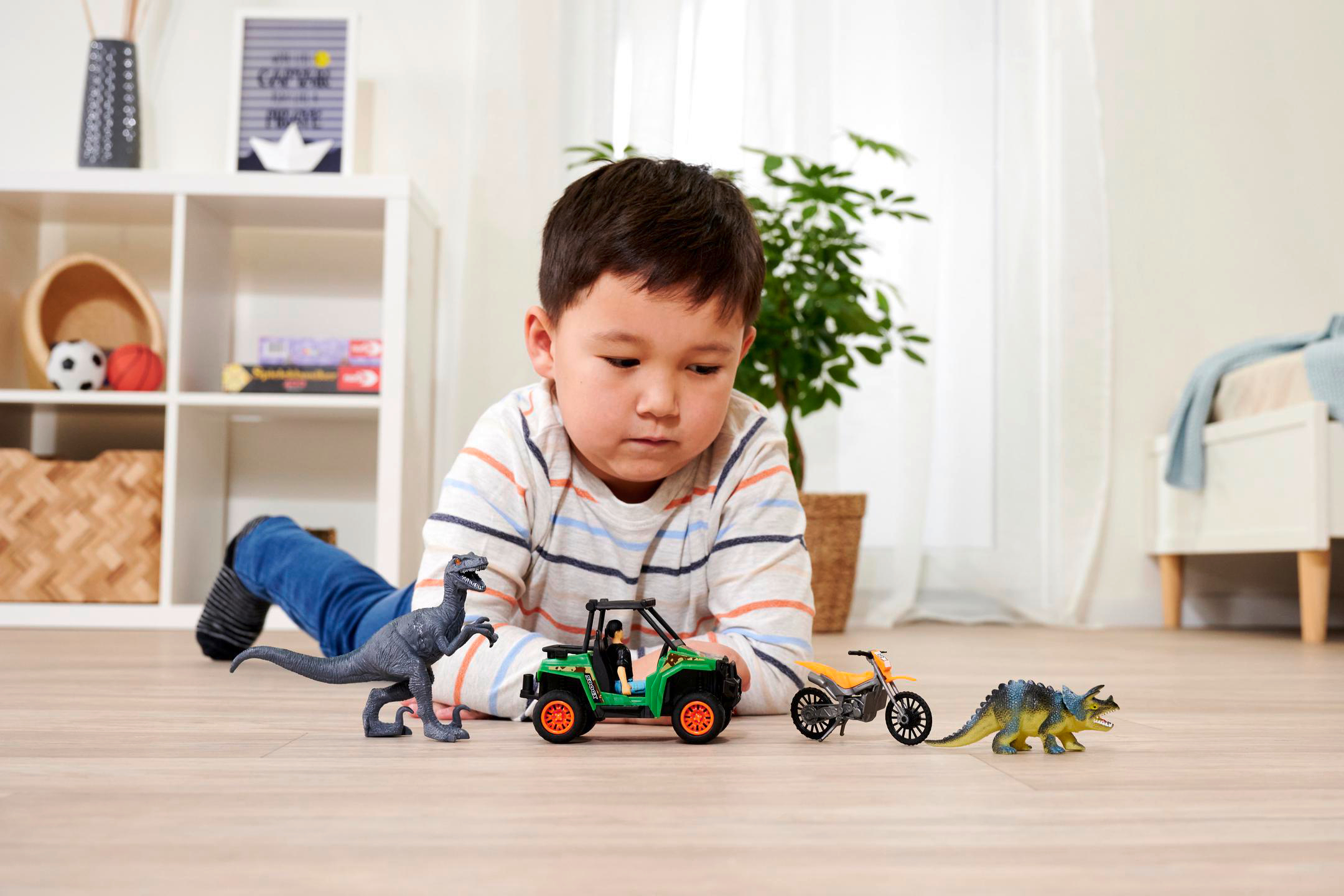 Junge spielt mit Spielzeug-Dinosauriern und Auto auf dem Boden. Weiße Regale im Hintergrund.