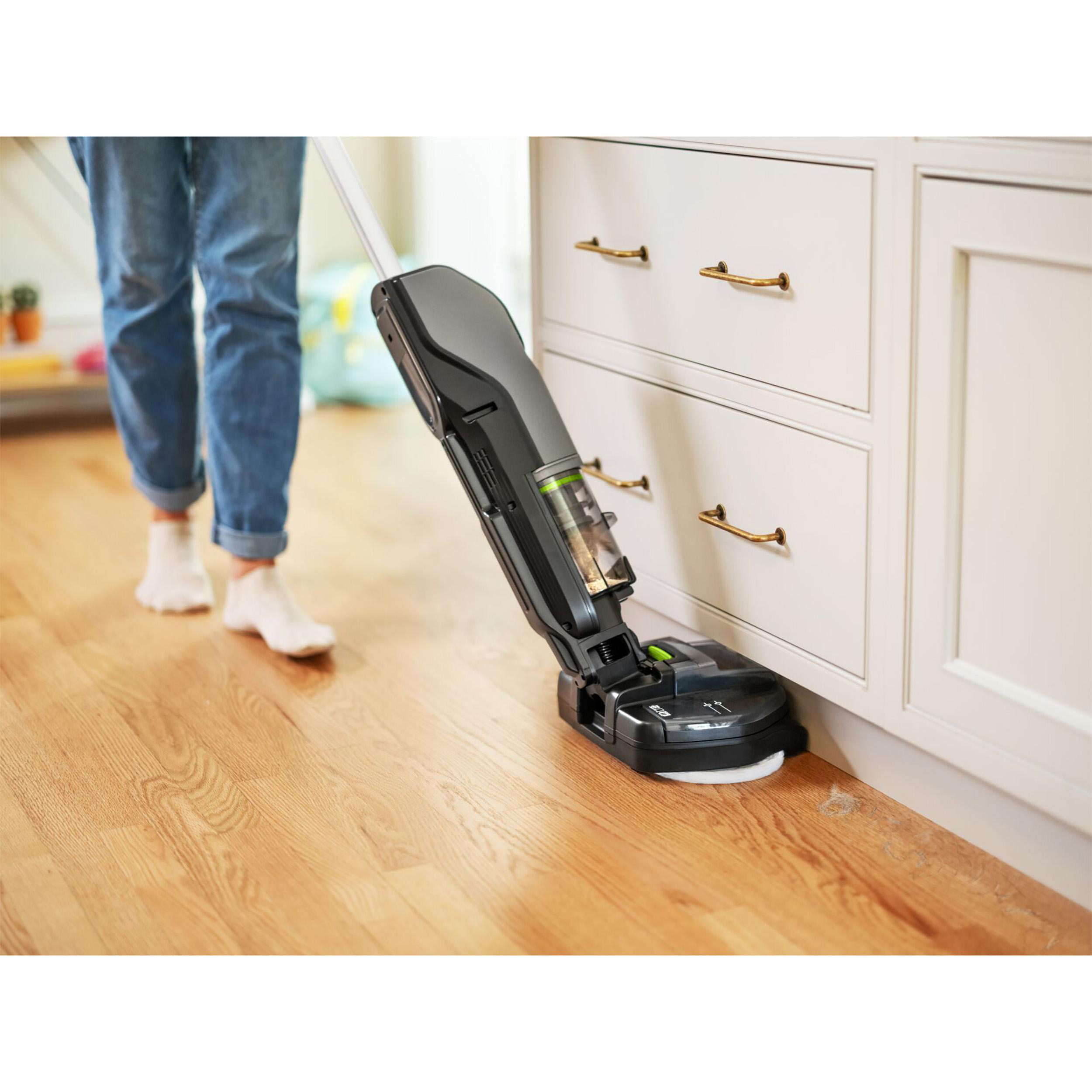 Eine Person benutzt einen schwarz-grauen Staubsauger auf einem Holzboden. Die Maschine steht in der Nähe eines weißen Schranks.