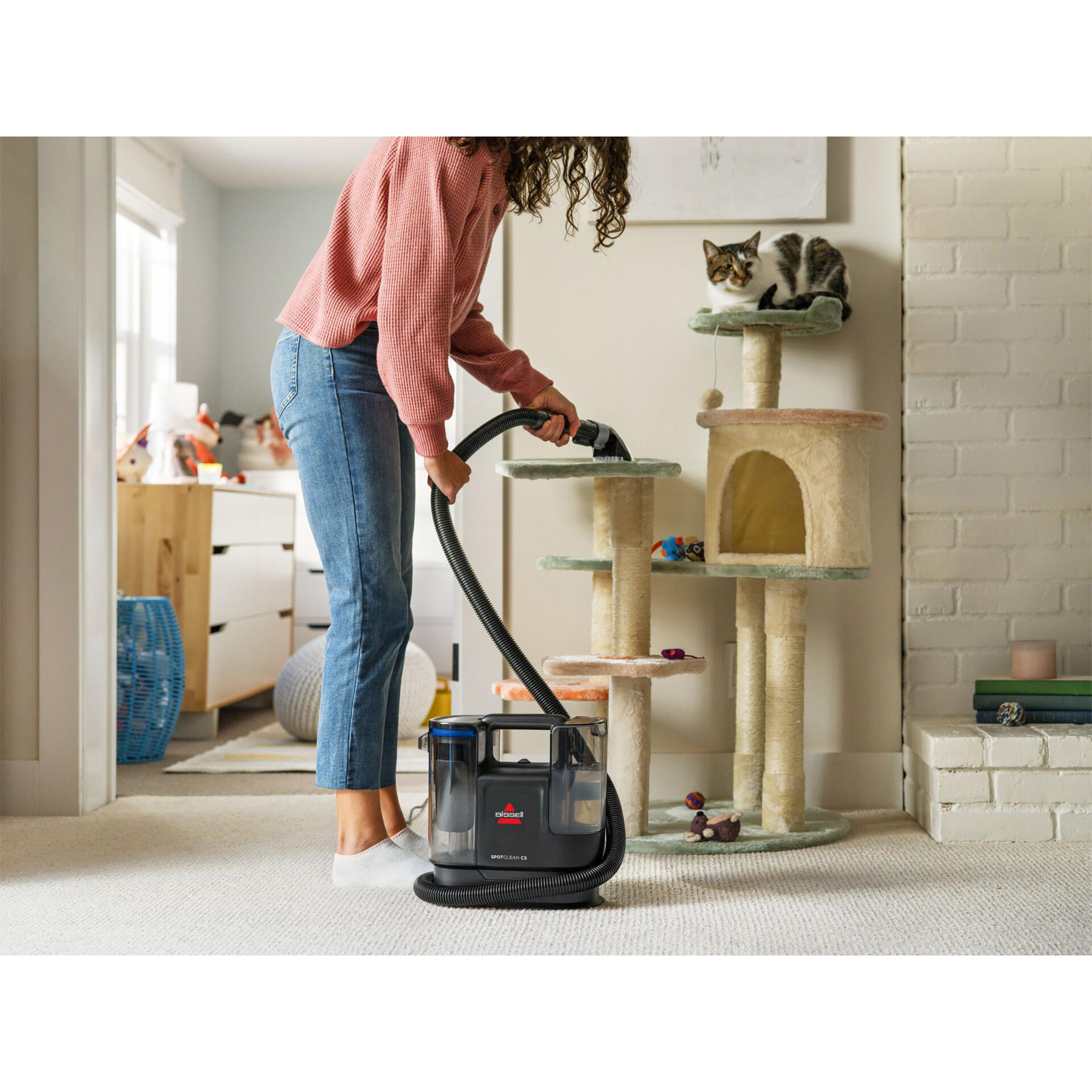 Eine Frau beim Staubsaugen, mit einem Katzenbaum und zwei Katzen im Hintergrund. Eine Reinigungsmaschine sichtbar.