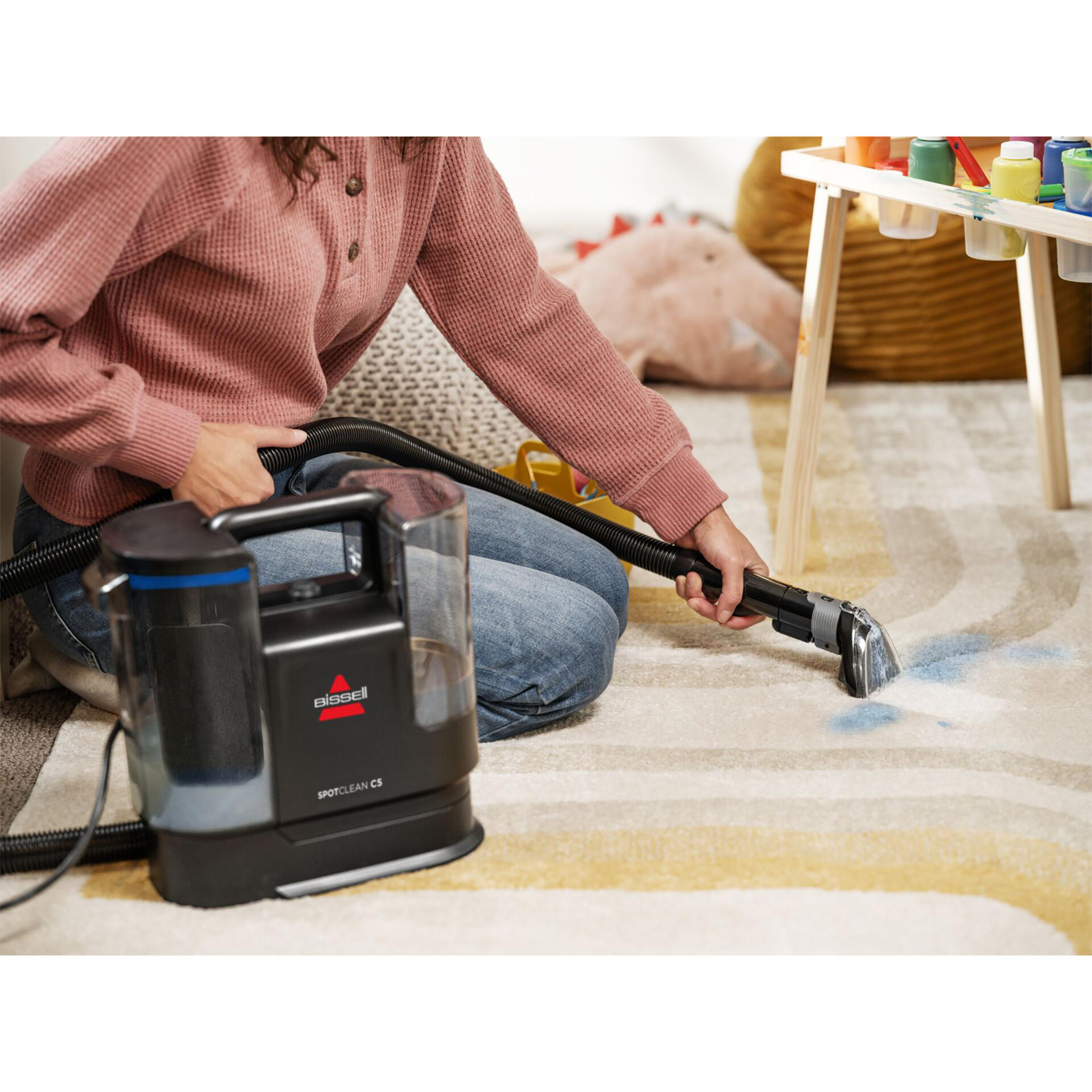 Eine Person saugt einen Teppich mit einem Bissell SpotClean. Die Maschine und ein Kinderspielbereich sind im Hintergrund.