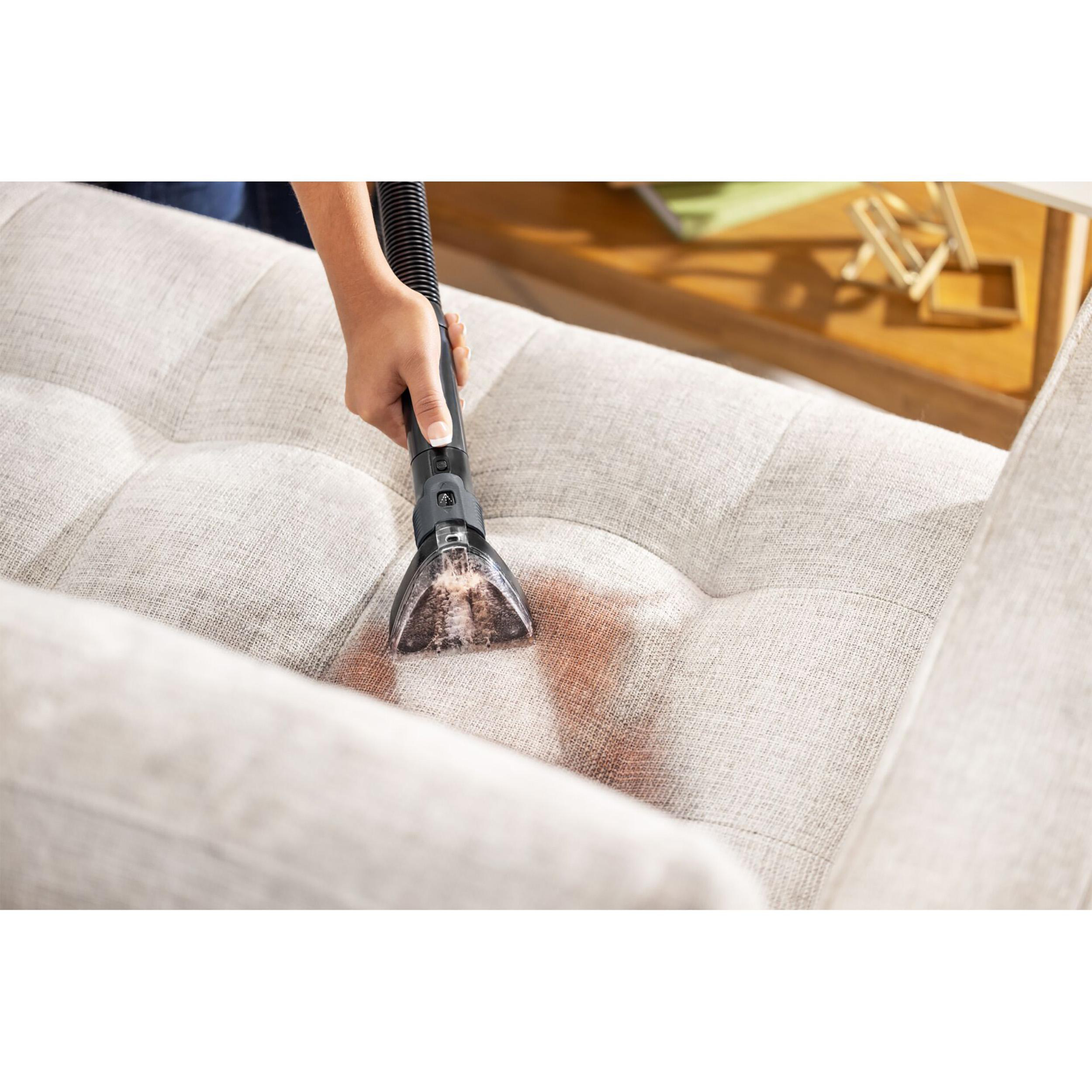 Eine Person saugt ein beiges Sofa mit einem Handstaubsauger ab. Das Sofa hat einige rote Flecken.