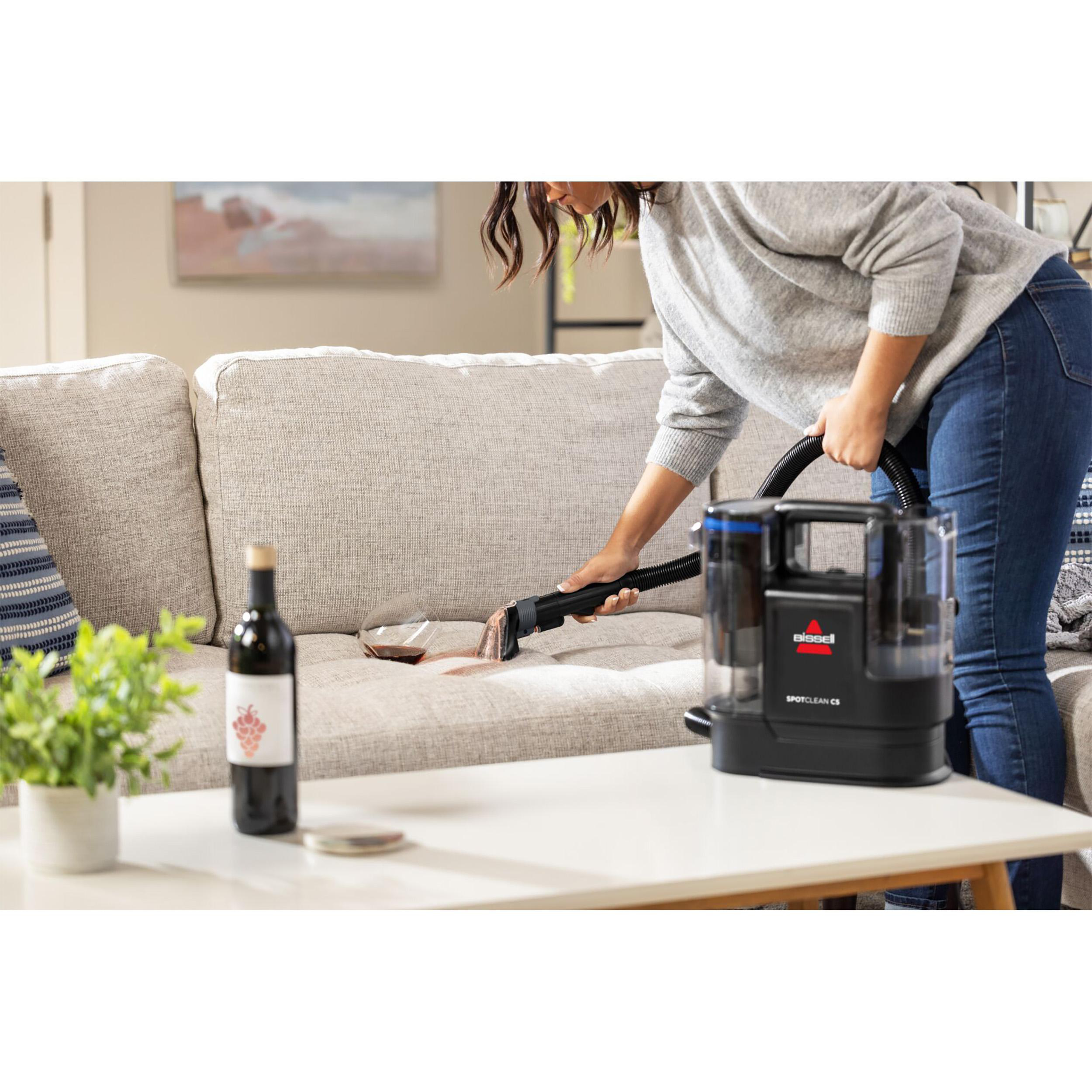 Frau reinigt Sofa mit Staubsauger. Weinflasche und Pflanze stehen auf dem Tisch.