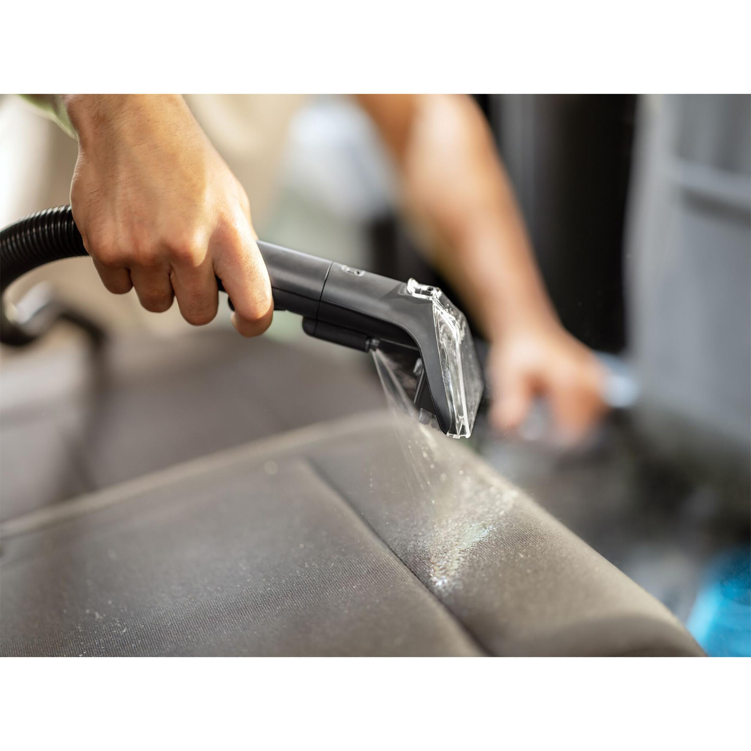 Person reinigt einen Autositz mit einem Reinigungswerkzeug, Wasser sprüht auf den Sitz, in einem Auto.