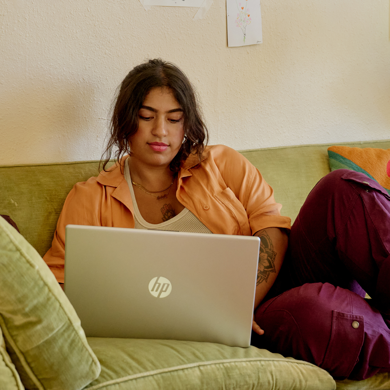 Woman is sitting on a green couch, using a laptop. She is wearing an orange top and purple pants.
