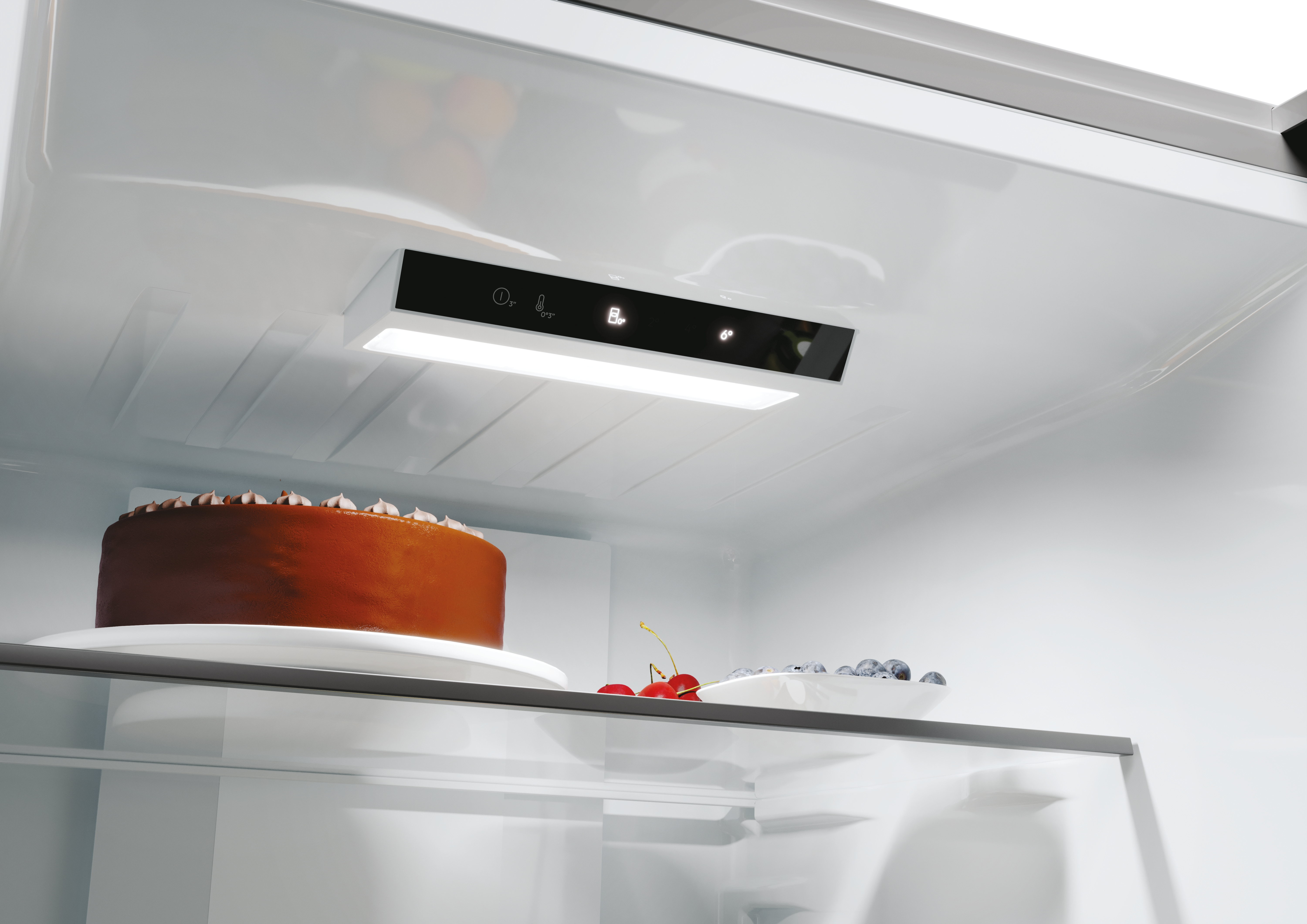 Interior de un refrigerador con estantes. Un pastel y fruta están en los estantes, con un panel de control arriba. Iluminación interior brillante.