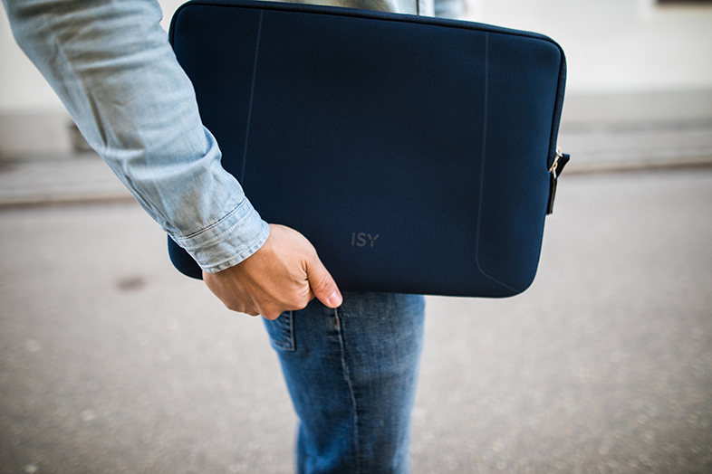 Személy sötétkék laptop tokot tart. Farmernadrág és világos farmering látható.