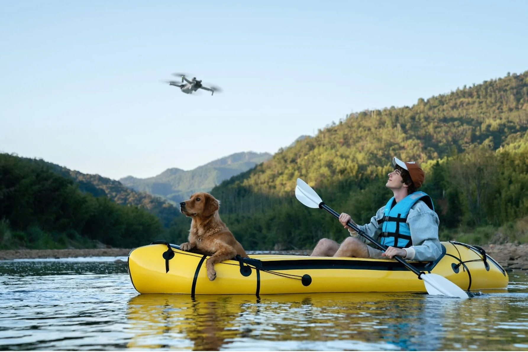 Mężczyzna i pies w żółtej kajaku na rzece. Dron leci nad nimi, góry w tle.