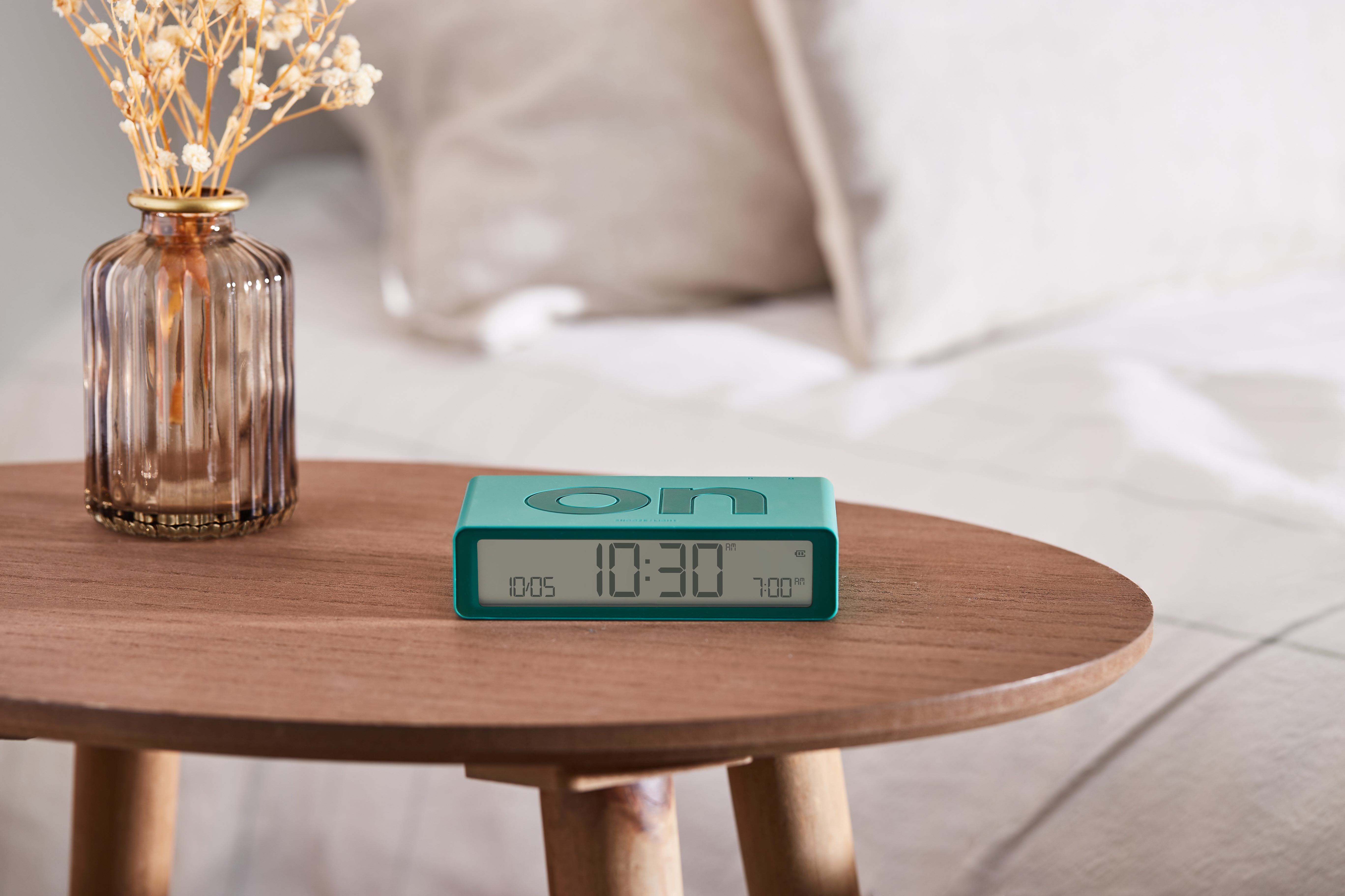 Sveglia turchese su un tavolo di legno. L'orologio segna le 10:30. Un vaso di fiori è vicino.