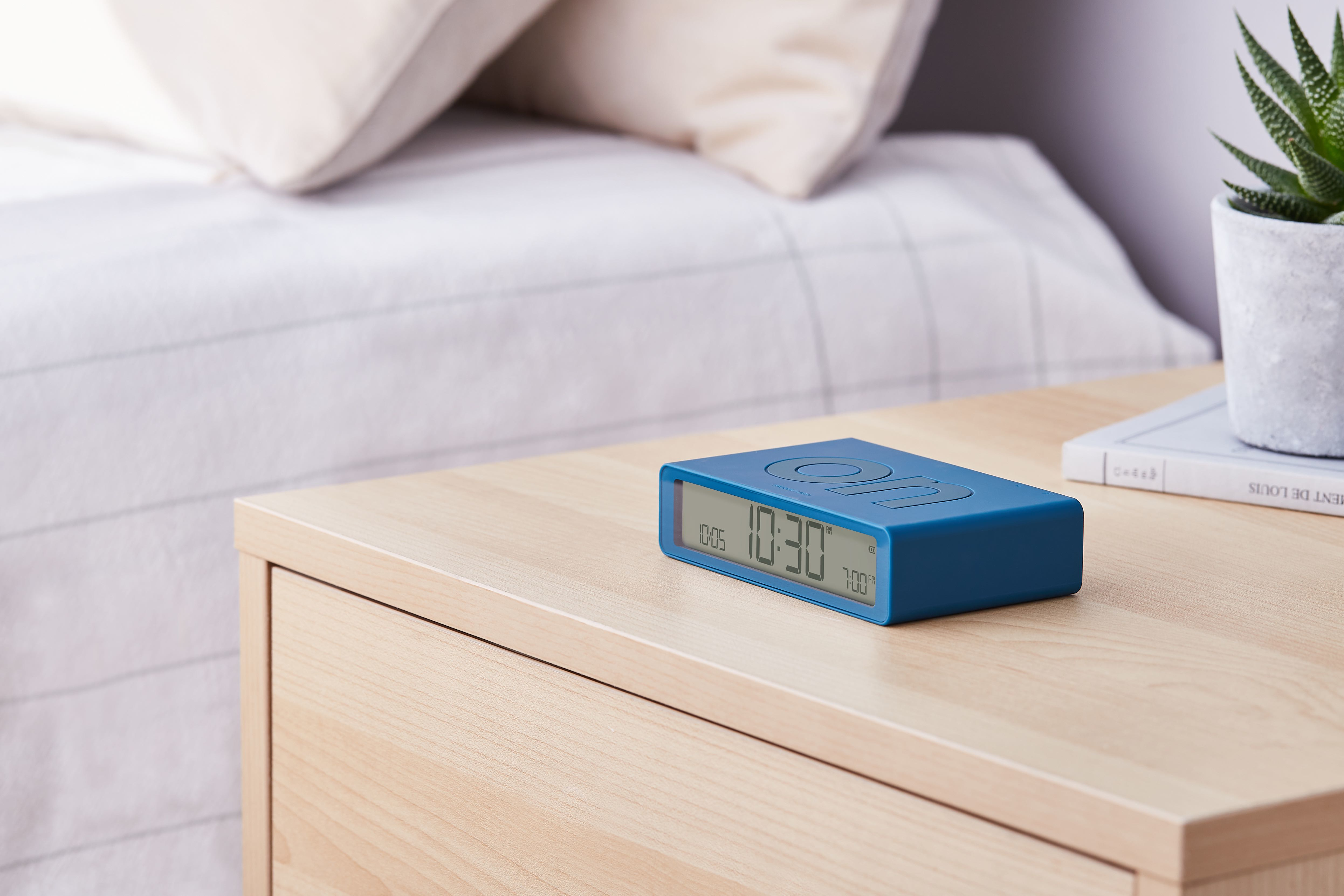 Sveglia digitale blu su un comodino in legno, con un libro e una pianta.