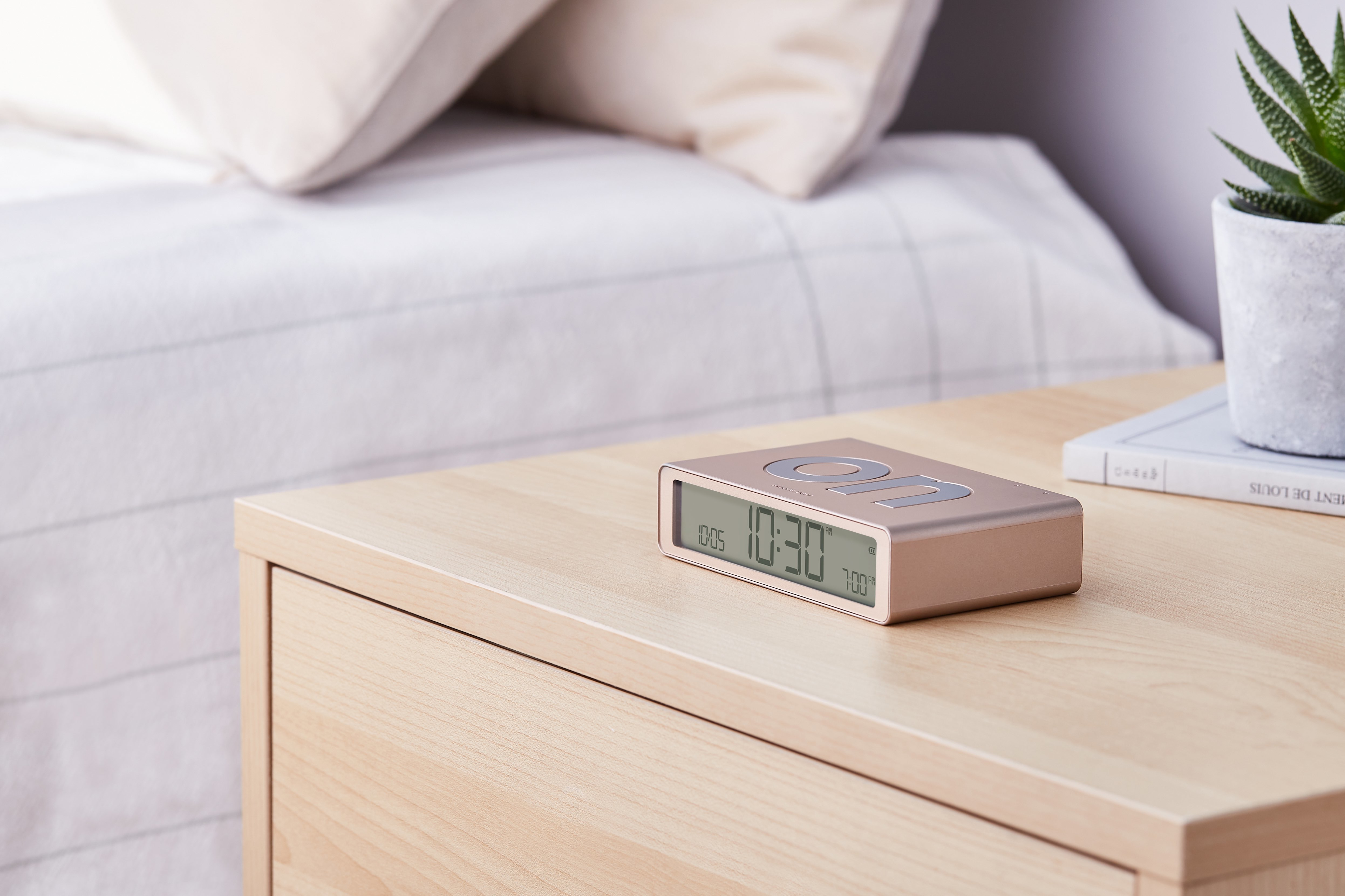 Una sveglia digitale dorata è su un comodino di legno vicino a un libro e una piccola pianta.