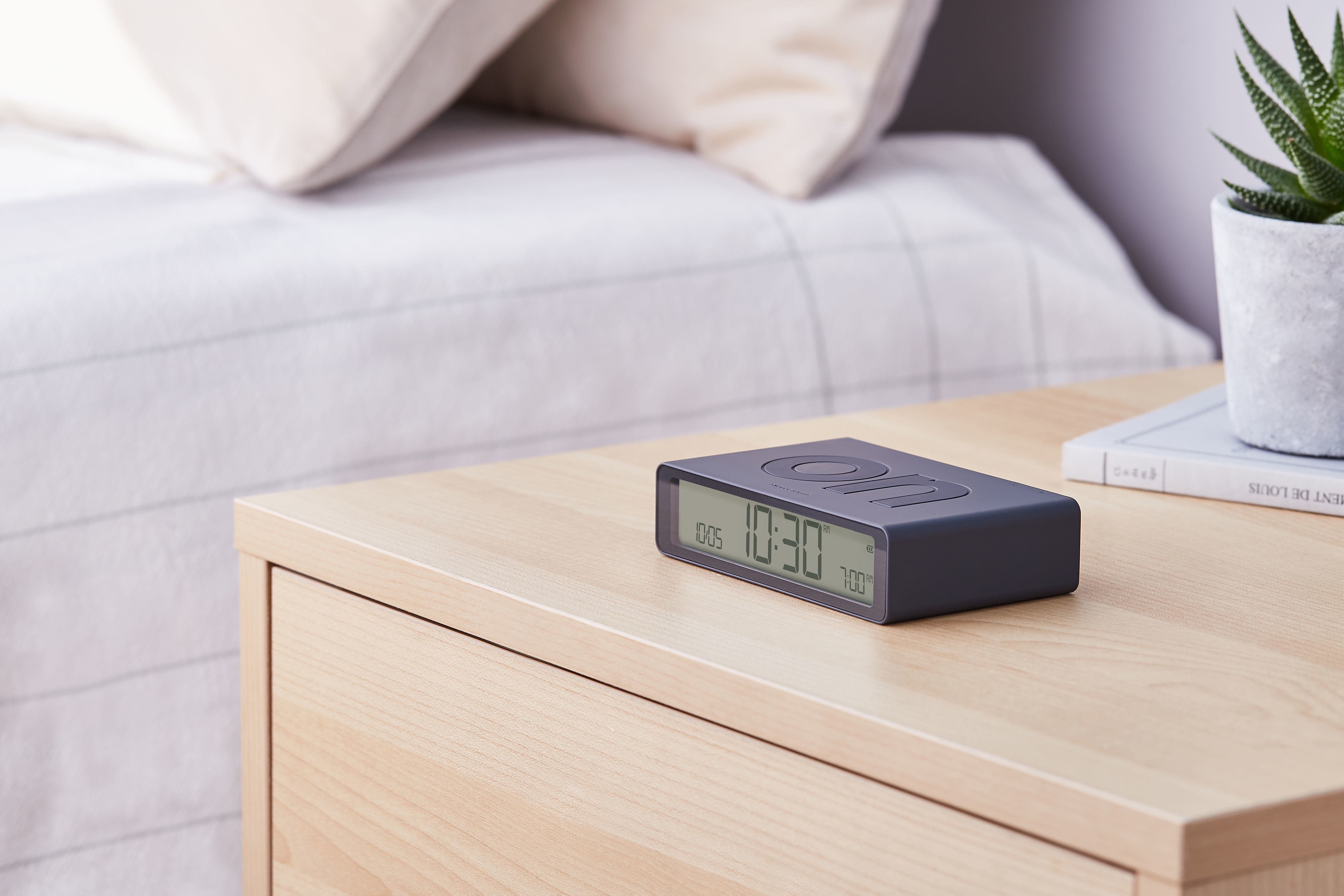Un orologio digitale su un comodino in legno mostra l'ora, con un letto sullo sfondo.