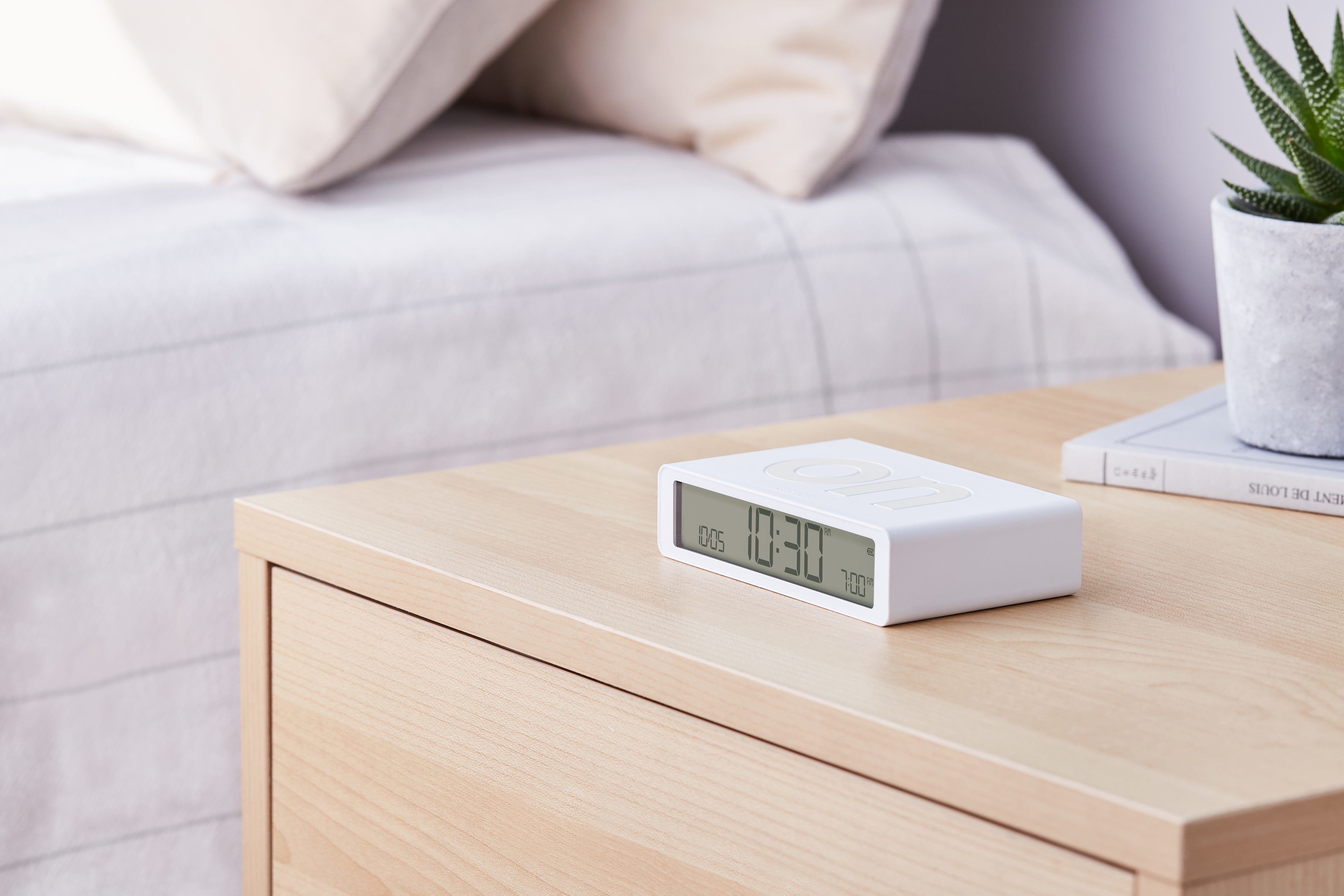 Weißer Digitalwecker auf einem Nachttisch aus Holz, Bett und Pflanze im Hintergrund.