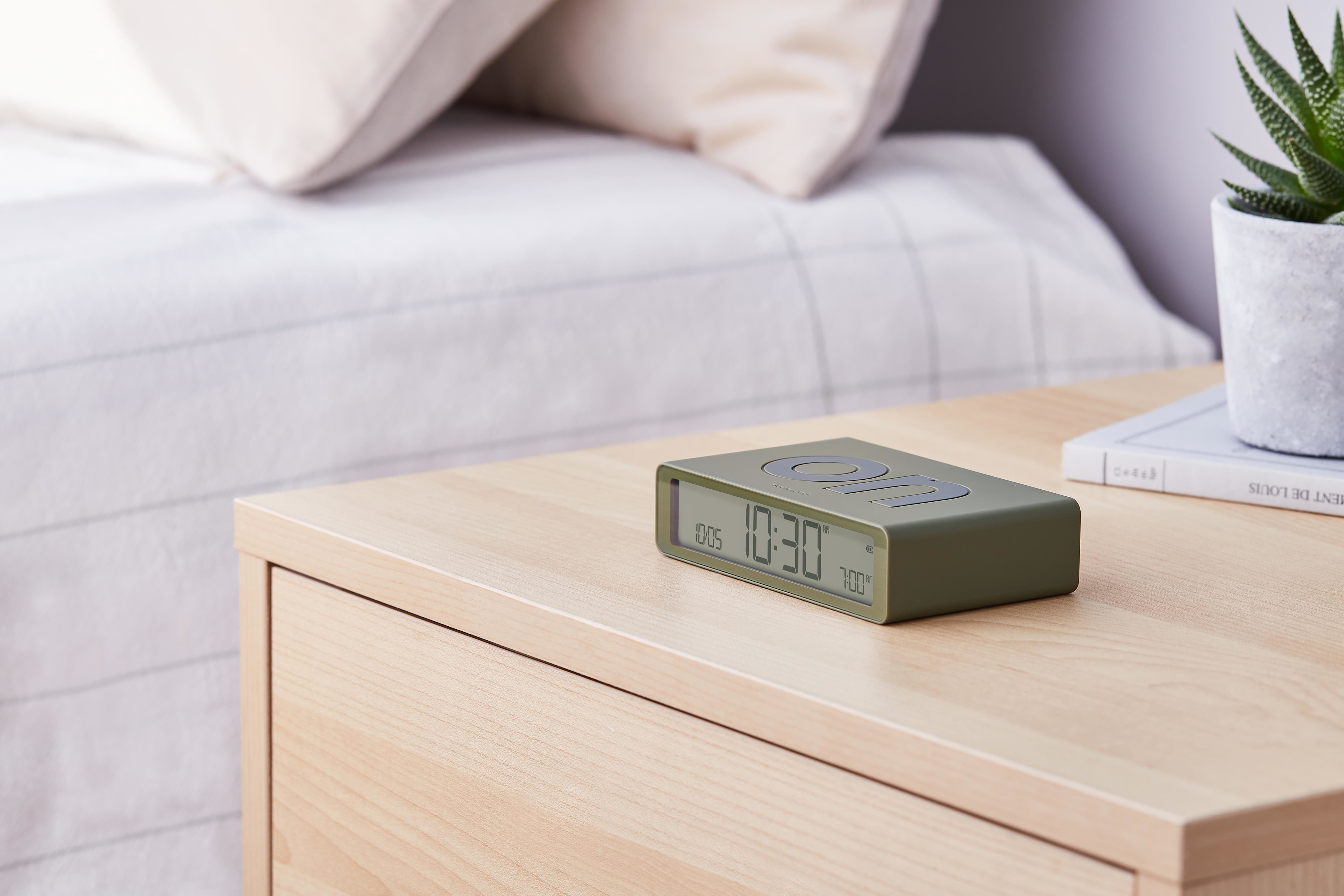 Orologio digitale verde su un comodino in legno accanto a una pianta e un letto con lenzuola bianche.