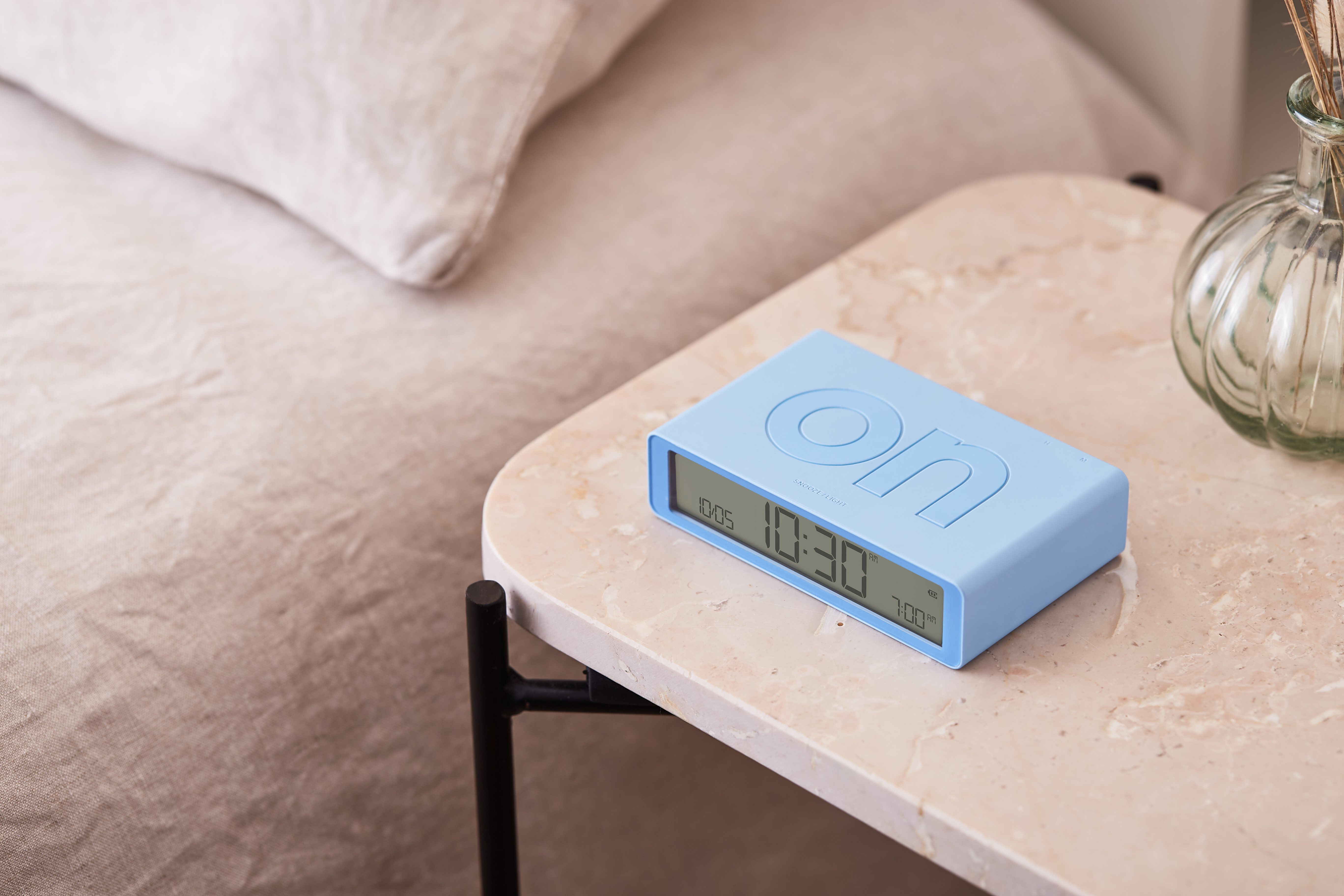Un orologio flip 'on' azzurro su un tavolo di marmo accanto a un letto e un vaso.