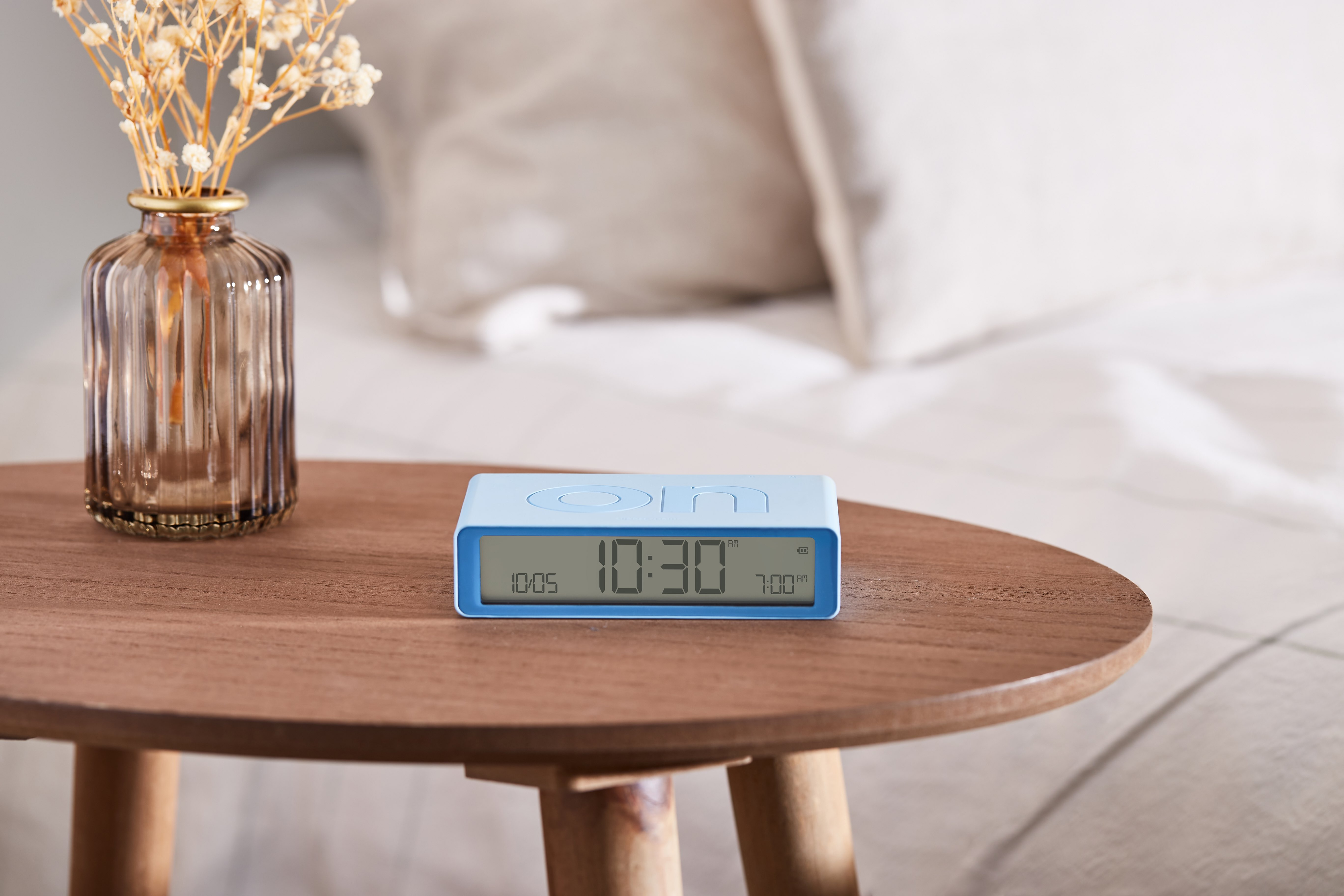Sveglia blu su un tavolo di legno con un vaso e letto sullo sfondo.