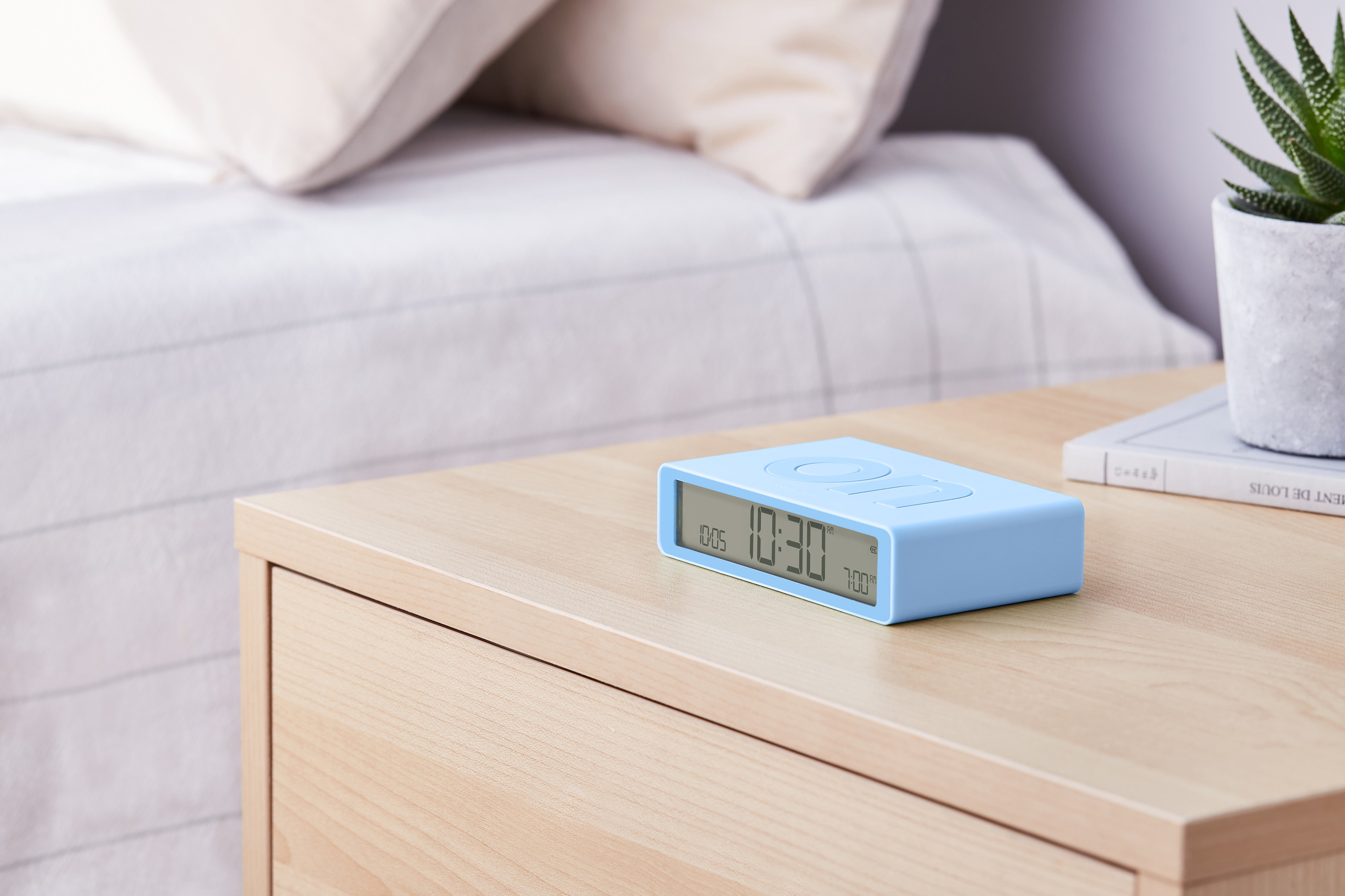 Orologio digitale blu su un comodino in legno vicino a un letto. Sfondo bianco.
