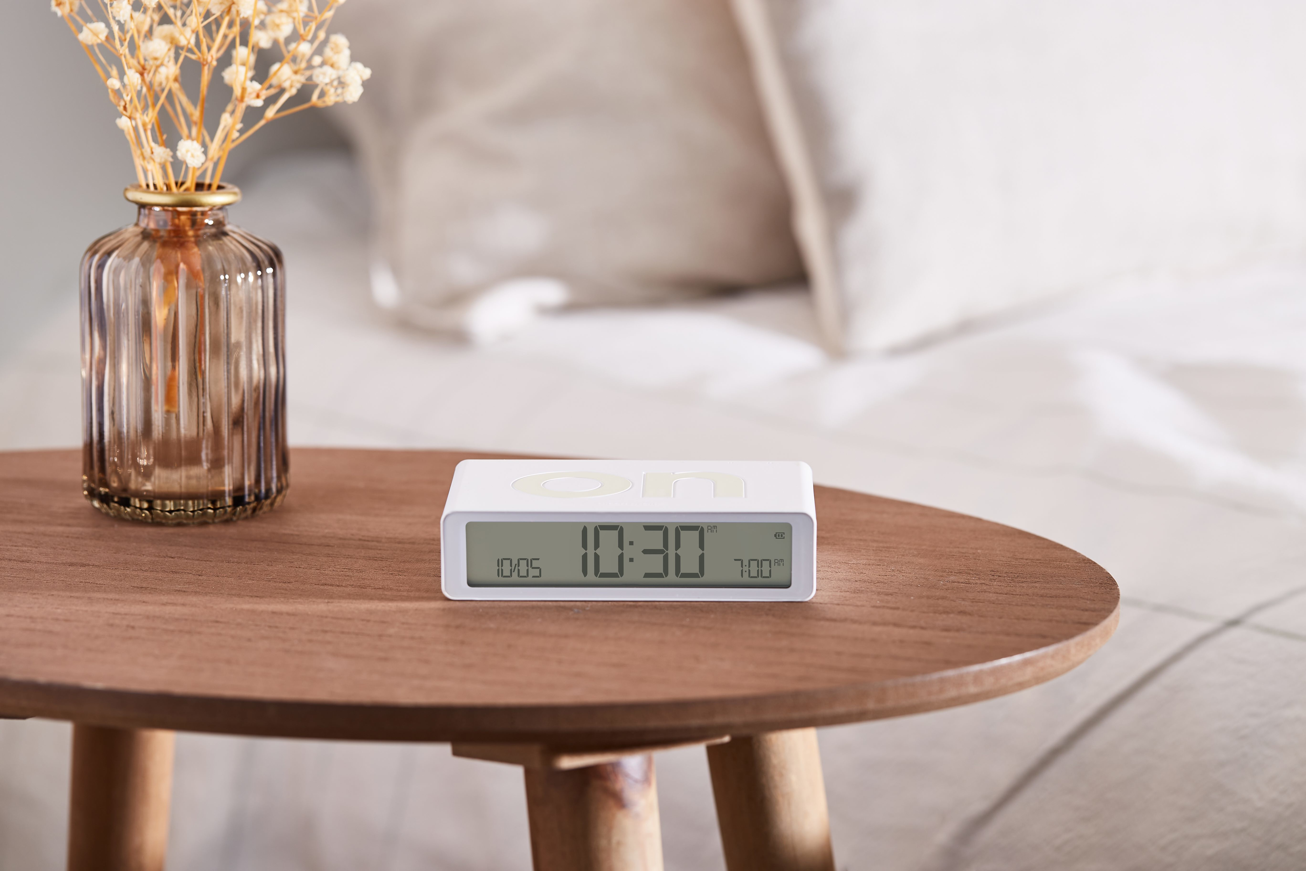 Eine weiße Digitaluhr auf einem Holztisch, mit einer Vase mit Blumen im Hintergrund.